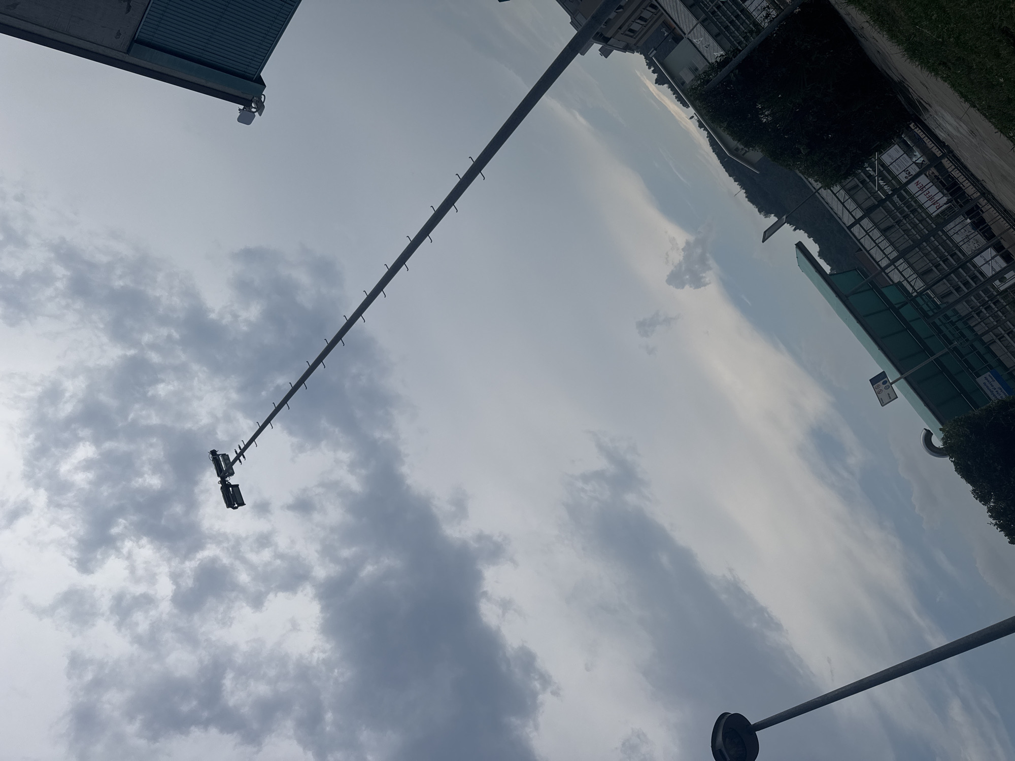 The image, captured in Chiasso, Switzerland, depicts an outdoor scene under an overcast sky. In the foreground, a tall, slender lamppost with a circular light fixture stands at an angle. Further into the scene, a much taller, thin pole with numerous short, horizontal attachments ascends diagonally towards the sky, topped with two floodlights. To the right, the corner of a building with a sloped, dark-colored roof and light blue corrugated siding is visible, also featuring two small lights mounted near the edge. In the middle ground, a structure with a green roof and a metal fence runs horizontally. Beyond this, a densely vegetated hillside rises, partially obscured by trees and buildings. The sky is filled with a variety of clouds, ranging from dark, heavy masses to lighter, wispy formations. The overall impression is of a slightly gloomy, daytime environment.