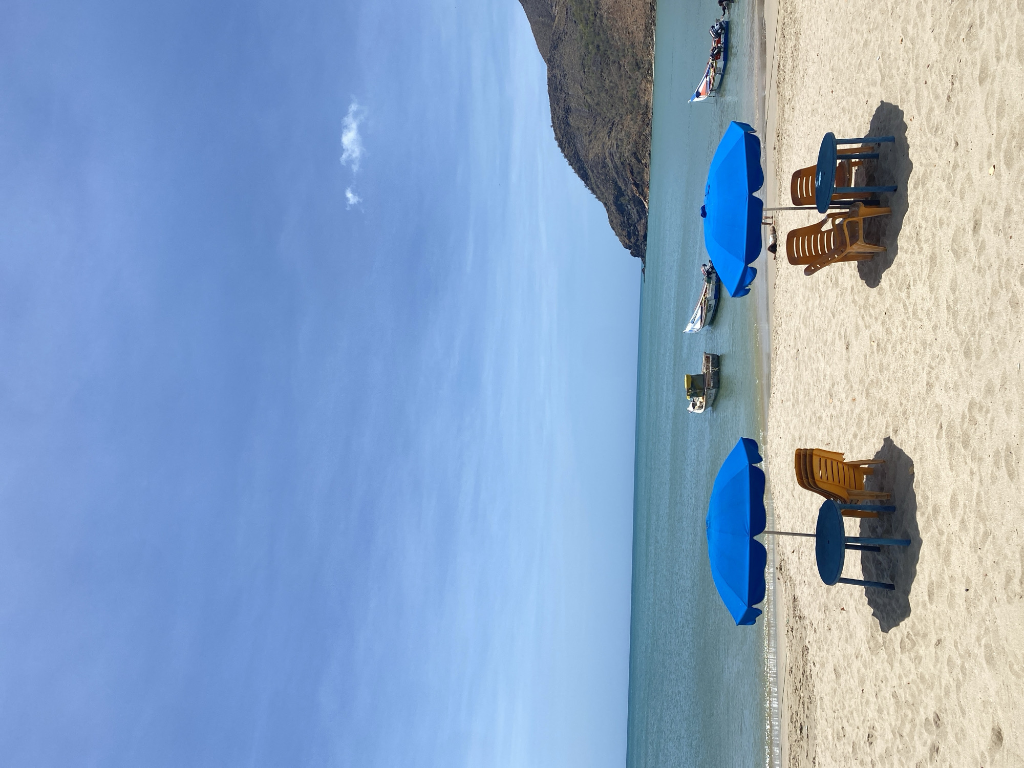 The image shows a serene beach scene, likely in Juan Griego, Venezuela, given the presence of a mountain extending to the sea. The focus is on two sets of beach furniture: blue umbrellas paired with blue tables and stacked yellow chairs. The sets are situated on a light tan sandy beach. The water is a pale green-blue hue, with small boats visible in the distance. A mountain range is located to the side of the furniture sets, with a bright blue sky above.
