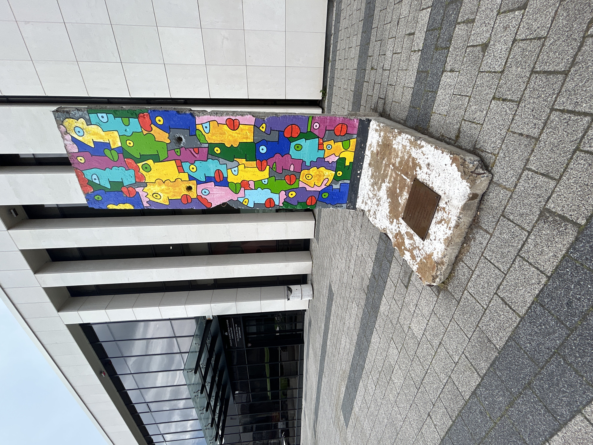 A vertical segment of the Berlin Wall, approximately 2.5 meters tall, stands upright on a roughly L-shaped concrete base in front of the City Gallery of Fine Art at 2008 Bulgaria Blvd, Plovdiv, Bulgaria. The wall segment is painted with a vibrant, pop-art style mural depicting numerous stylized, interlocking human faces in shades of yellow, green, blue, pink, and purple, each with distinct circular eyes and red lips. Several small, circular holes are visible within the painted faces. A rectangular bronze plaque is affixed to the weathered concrete base.
The wall is situated on a paved outdoor area composed of light grey rectangular pavers, interspersed with darker grey strips, leading towards a modern building. This building features a light-colored, possibly stone or concrete, facade with prominent vertical columns and large, dark, reflective glass window panels. A clear glass canopy extends over the main entrance on the left side of the frame. A sign near the entrance, visible in both Bulgarian and English, identifies the location as "CITY GALLERY OF FINE ART 2008 Bulgaria Blvd." A white cylindrical waste receptacle is positioned to the right of the entrance. The scene is illuminated by an overcast sky, with no individuals present.