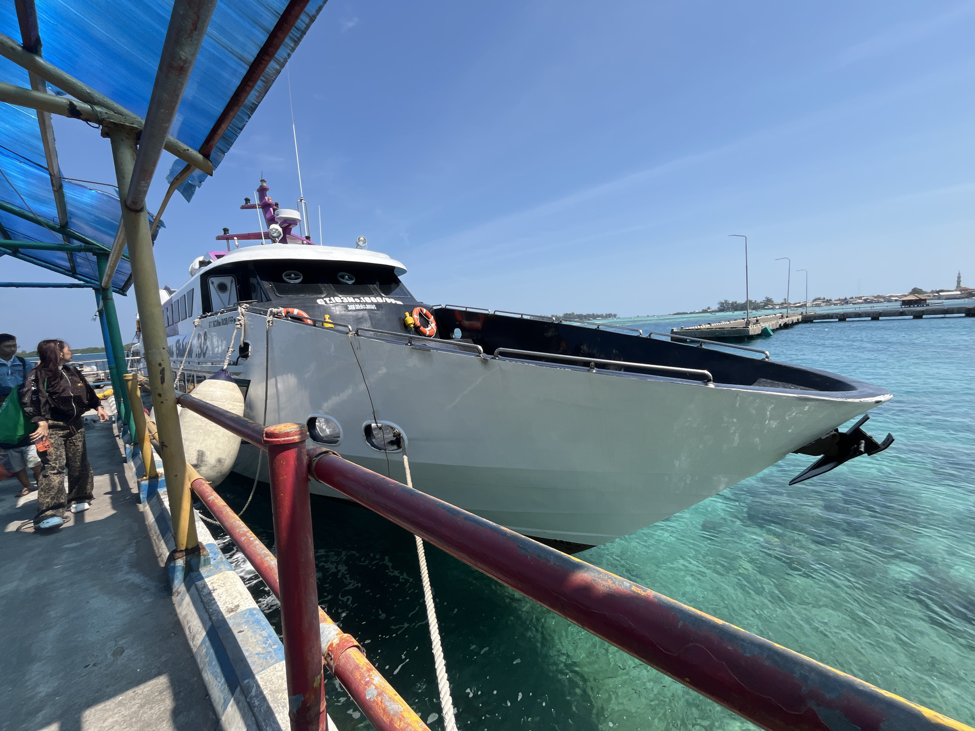 In Karimunjawa, Indonesia, a white passenger ferry is docked at a pier under a blue canopy. The ferry, named "ET:ICAKAN.1008/89," has a black hull line and a purple radar array. Two orange life preservers are visible on its side. Three people stand on the pier to the left of the ferry, and one is walking away from the camera. The water is a clear turquoise, and a pier with streetlights extends into the distance. The sky is bright blue with scattered clouds.