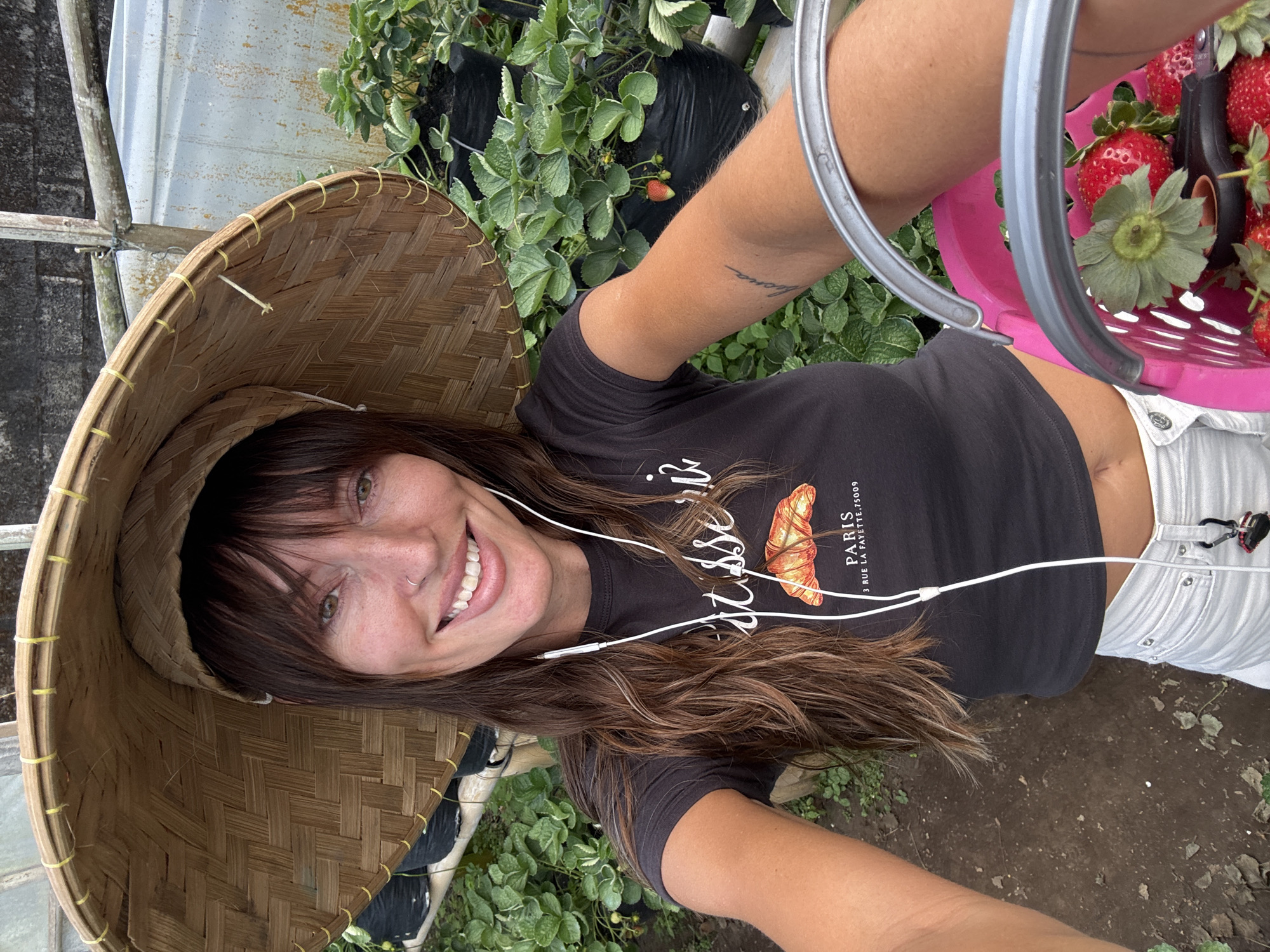 In Sukasada, Indonesia, a woman with long brown hair, wearing a large conical straw hat, a dark t-shirt with a croissant graphic, and white jeans, smiles at the camera. She has earphones in her ears and a small nose ring. She is in a greenhouse or outdoor farm setting with rows of strawberry plants in black bags visible in the background. In her right hand, she holds a pink basket filled with freshly picked strawberries and pruning shears. The ground is covered in dark soil with some scattered leaves.