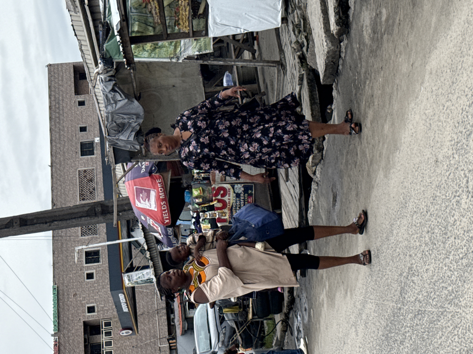 A medium shot captures two women and a toddler on a paved street in Ikorodu, Nigeria. The woman on the left, holding the toddler, wears a tan, off-the-shoulder dress with a colorful neckline and black leggings. Her hair is braided, and she carries a blue tote bag. The toddler is held in her left arm, wearing a light-colored shirt. The woman on the right is dressed in a black floral-print dress and black sandals. The environment includes a multi-story building with a sign reading "SUPERMARKET" in green letters, and an "AUTO ROAD" gas station sign. In the foreground, there are market stalls with awnings, stacks of tires, and weathered concrete slabs. The sky is overcast.