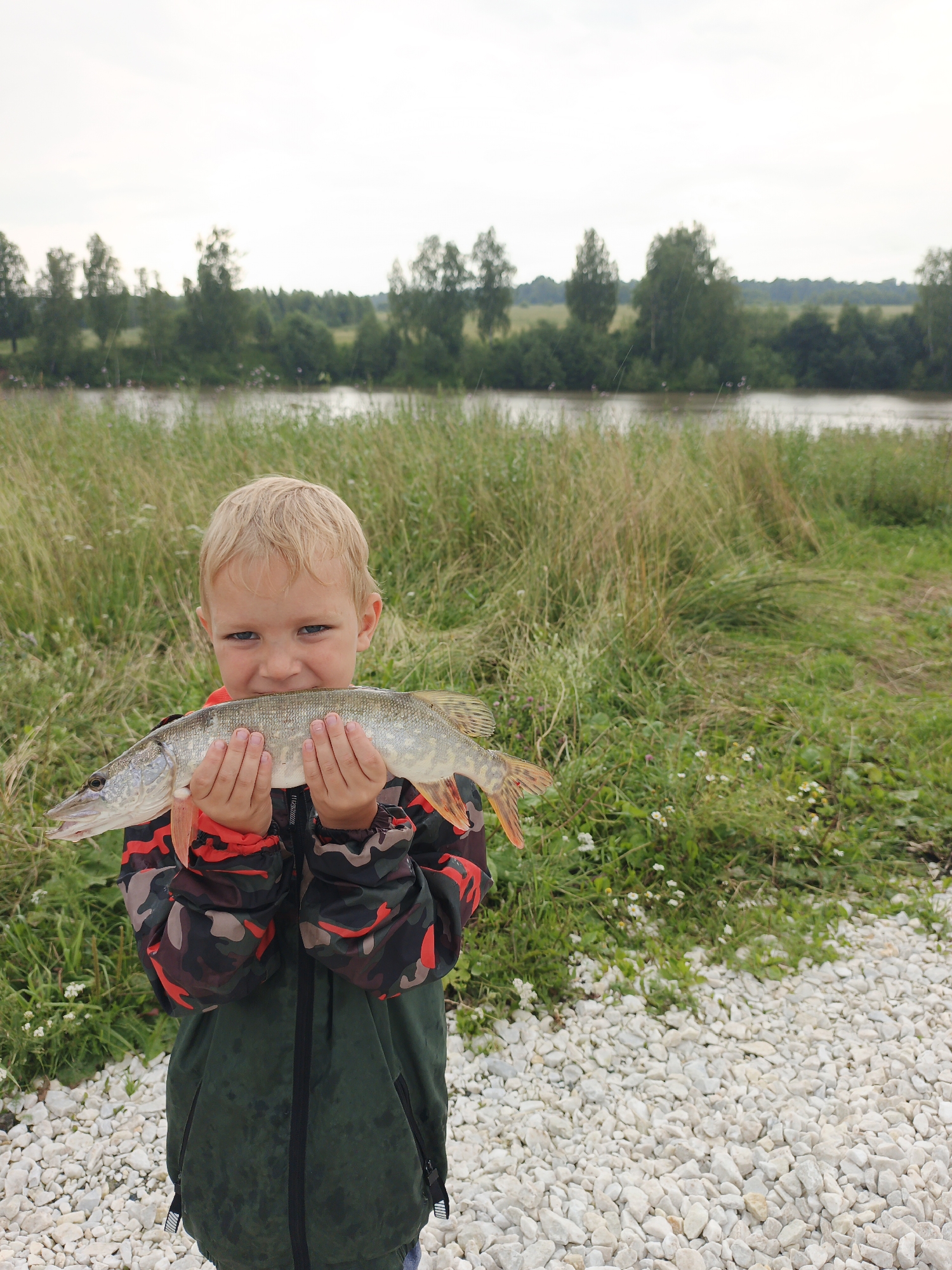 In the city of Urtiushka, Russia, a young boy with blond hair and blue eyes stands outdoors holding a freshly caught pike. He is wearing a camouflage jacket with red accents. The background shows a body of water, tall grass, trees, and a cloudy sky. The boy is positioned in the foreground, and the fish is held horizontally in front of him. The ground in front of him is covered with white gravel.