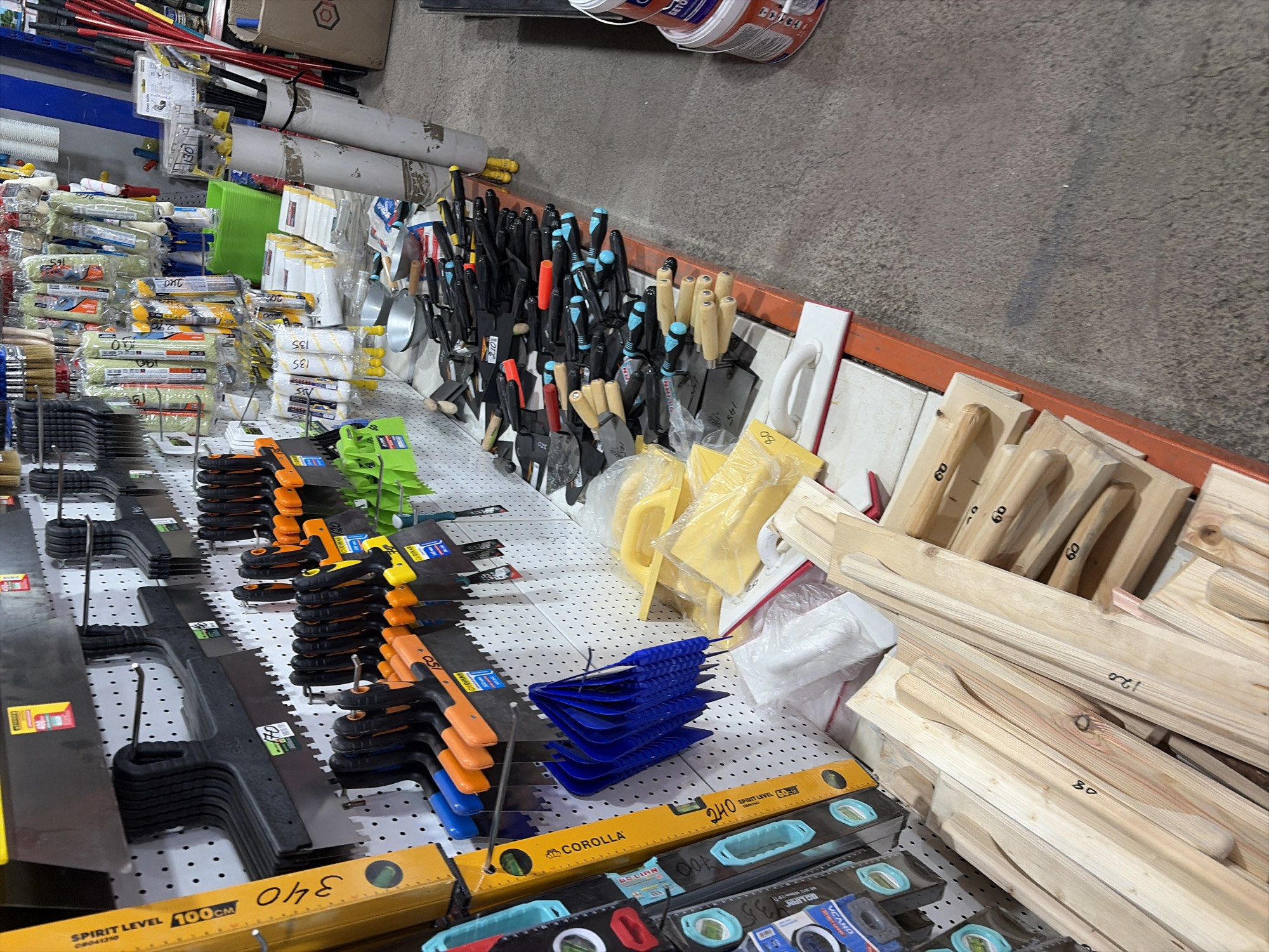 The image shows a retail display of construction tools and materials. The items are arranged on pegboards and shelves. Visible items include trowels, spatulas, levels, paint rollers, brushes, and wooden planks. The tools have various handle colors, including black, orange, and blue. Several items have price tags or labels. The background appears to be a store interior with concrete flooring. There are no people visible in the image. The scene is a static display of merchandise, typical of a hardware or building supply store. The location is identified as being in Kant, Kyrgyzstan.