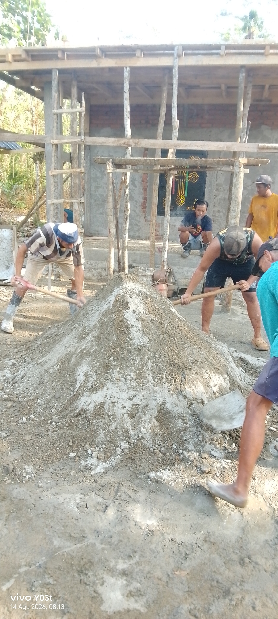 In Woiha, Indonesia, at a construction site, several men are actively engaged in mixing cement. The scene is dominated by a large mound of sand and gravel, positioned in the foreground. To the left, a man in a striped shirt and light shorts is using a shovel to mix the pile, wearing a white and blue cap and tall boots. To the right of the mound, another man, wearing a camouflage tank top and dark shorts, is also using a shovel to mix the cement. In the background, a partially constructed building with wooden scaffolding and concrete walls is visible. A man in a yellow shirt stands to the far right, observing the work. Further back, a man in a blue t-shirt is crouched down, smoking and looking at his phone, with a decorative cross visible behind him. The ground is dusty and appears to be a mix of dirt and some wet cement. The lighting suggests it is daytime, likely late morning or early afternoon.