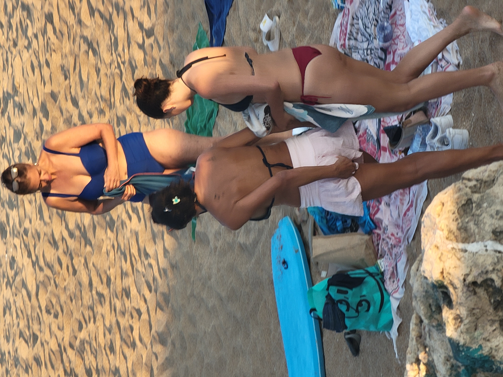 A group of three women are on a sandy beach in Quarteira, Portugal, near some rocks. The woman in the background, wearing sunglasses and a blue bikini, is holding a teal towel. To her right, another woman in a black bikini is holding a towel. In the foreground, a third woman in a black bikini and a light pink skirt is walking away from the camera. A blue surfboard is visible on the sand behind the women, along with a green bag with a face design, a brown paper bag, and towels spread on the sand. Two pairs of white sneakers are also on the sand. The lighting suggests it is late afternoon.