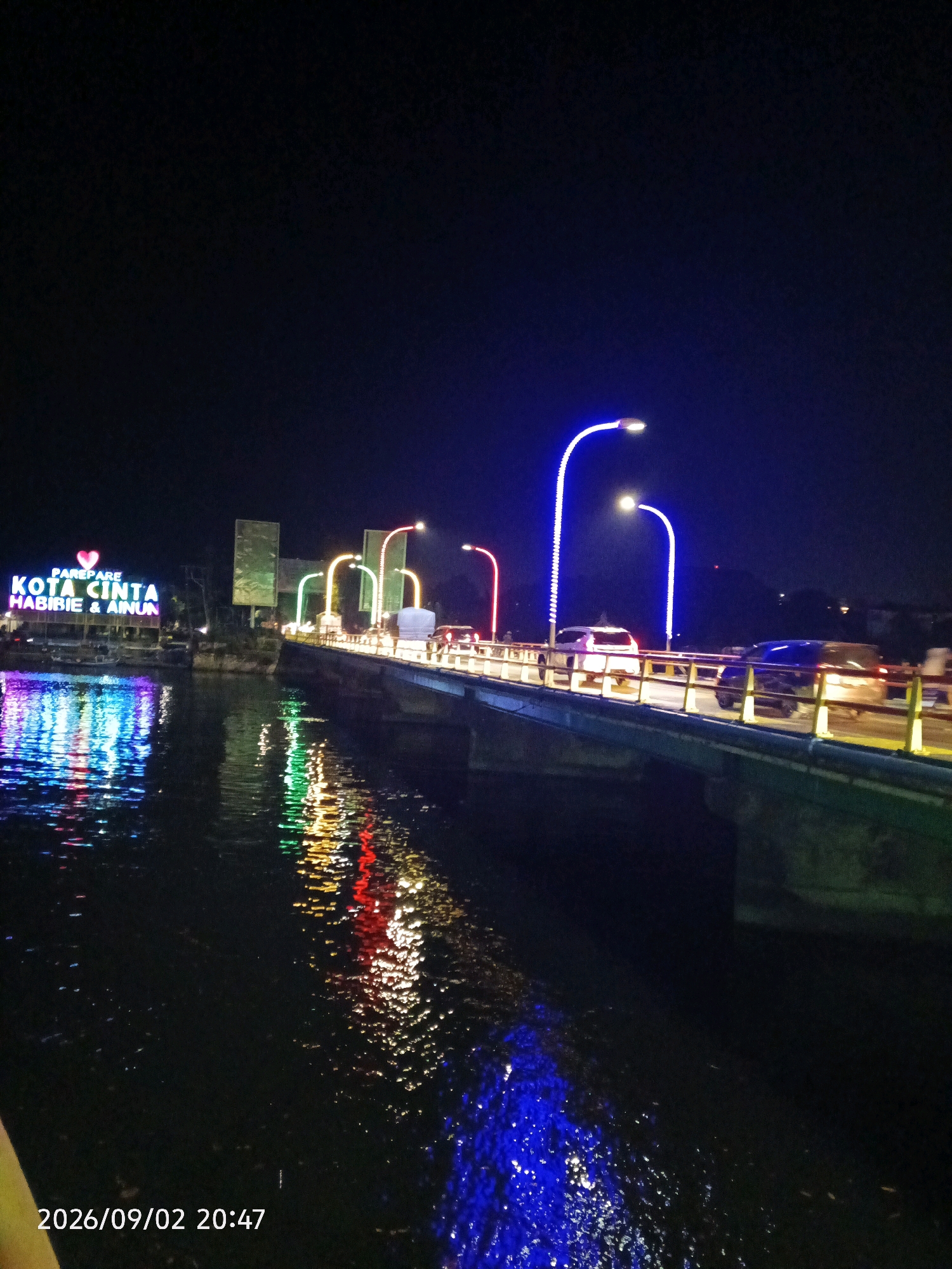 The image captures a nighttime view of the "Tugu Cinta Habibie & Ainun" (Habibie & Ainun Love Monument) and the adjacent bridge in Parepare, Indonesia. The monument, illuminated with vibrant neon lights in blue, purple, and pink, stands on the left side of the frame, its reflection shimmering in the dark water below. The bridge, which is also lit with colorful lights along its railings and street lamps, spans across the water. Cars are visible driving on the bridge, their headlights and taillights creating streaks of light. The water surface is disturbed by ripples, further reflecting the colorful lights from the monument and bridge. The date and time "2026/09/02 20:47" are overlaid on the bottom left corner of the image. The overall scene is one of a lively urban nighttime setting.