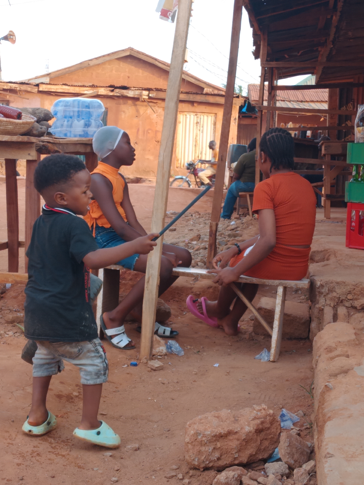 In Yelwa, Nigeria, a young boy in a black shirt and patterned shorts stands on a dirt ground. He holds a dark stick, pointing it toward two girls seated on wooden benches. The girl on the left wears an orange top and blue shorts with a white head covering. The other girl has her back to the viewer and wears an orange dress. The setting appears to be an outdoor marketplace or roadside area. In the background, there are buildings and other individuals. Various items are placed on wooden surfaces and shelves, potentially for sale.