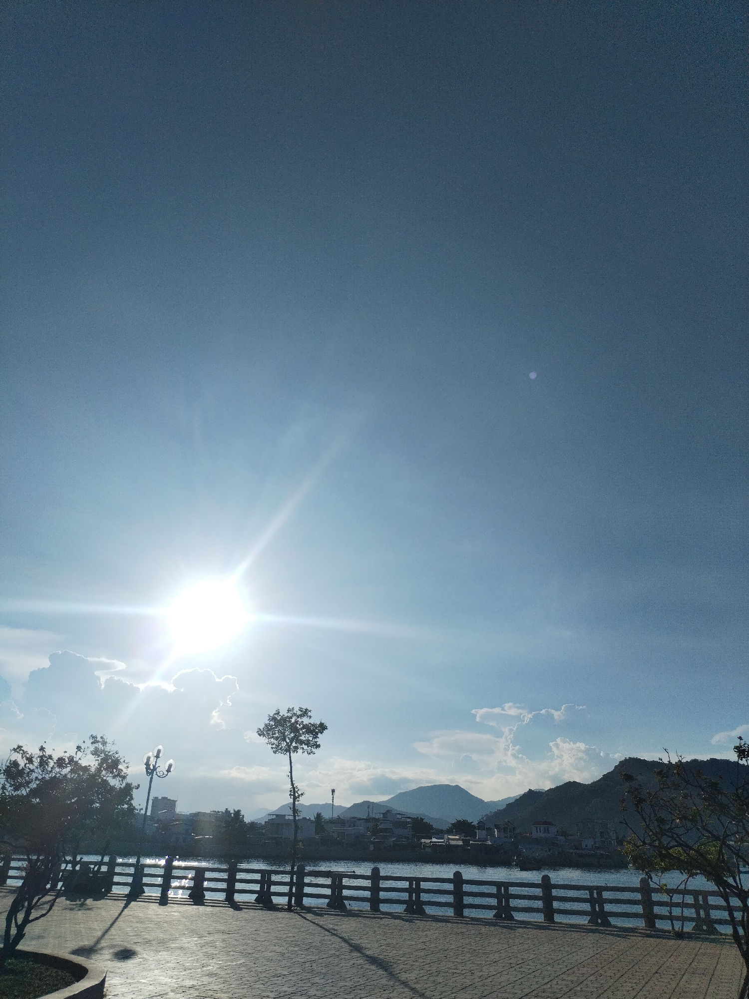 The image depicts a bright, sunny day in what appears to be a park or waterfront area in Nha Trang Ward, Vietnam. The sun is prominent and causes glare reflecting off the water. A low wooden fence runs along the waterfront. Trees are scattered along the edge of the frame. Buildings and hills are visible in the background. The foreground shows a paved area with a discernible pattern. The sky is blue with some clouds.