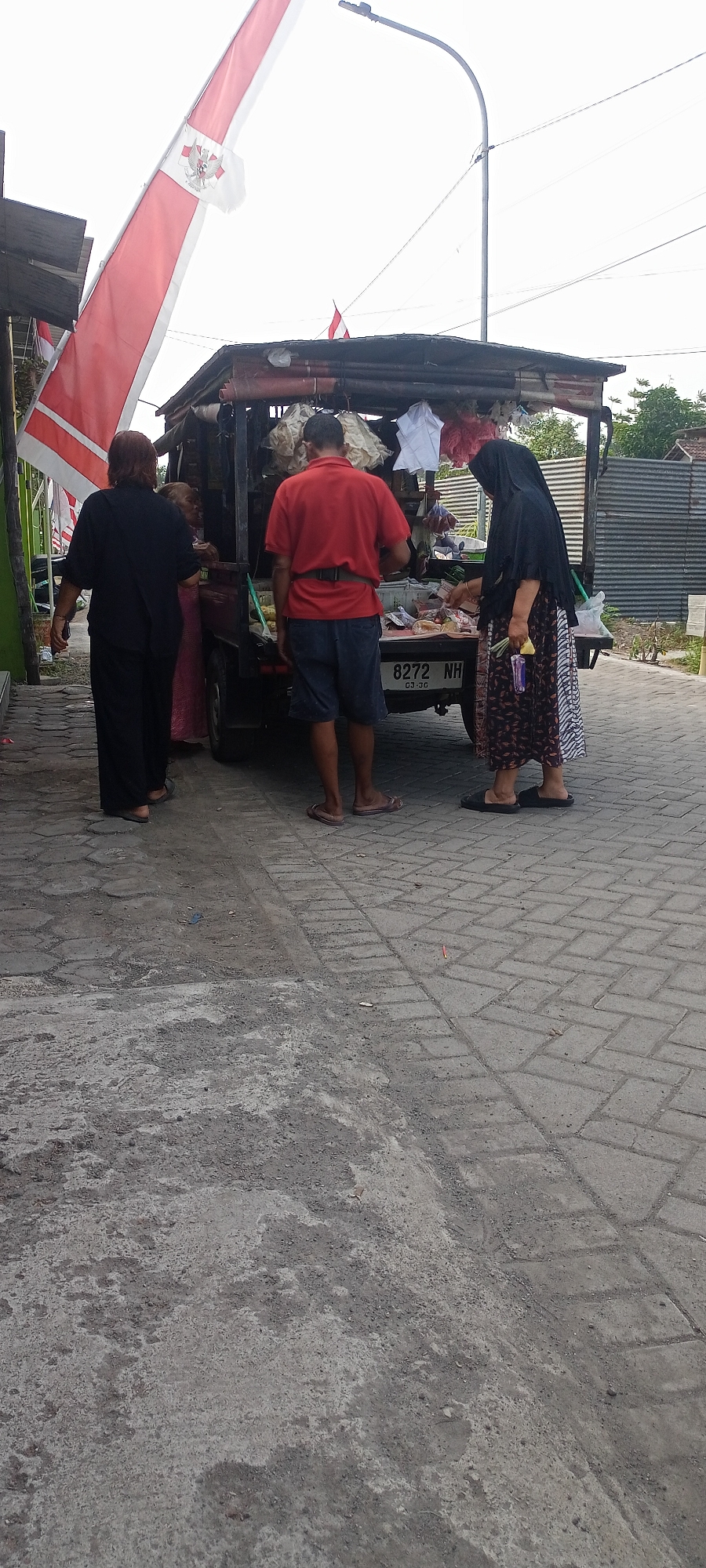 In Gempolklutuk, Indonesia, a scene unfolds by a roadside. The foreground shows a paved area with a distinct pattern of interlocking grey bricks and patches of rough concrete. A large red and white flag with an emblem, possibly a national flag, is prominently displayed to the left, fluttering against a pale sky. To the right, a tall, curved street light stands out, its base obscured by a wooden structure.
The central focus is a small, open-back truck serving as a vendor's stall. The truck bed is laden with various items, including what appear to be bags of goods and possibly food products displayed on a surface at the rear. Hanging from the top of the stall are several white and pinkish items, perhaps fabric or decorative elements. A small Indonesian flag is visible on the top of the stall.
Three individuals are interacting with the vendor. A woman dressed in black, with dark hair visible, stands to the far left, facing away from the viewer. Beside her, a woman in a pink garment is partially visible. A man in a red polo shirt and denim shorts stands on the right side of the truck, facing towards it, with a dark belt cinched around his waist. Another person, a woman wearing a black hijab and a patterned skirt, is interacting with the vendor at the back of the truck, holding what appears to be a bunch of green herbs or stalks. The license plate of the truck reads "8272 NH". The background shows a corrugated metal fence and some greenery, suggesting a residential or commercial area. The overall impression is of a bustling street-side market or roadside stall.