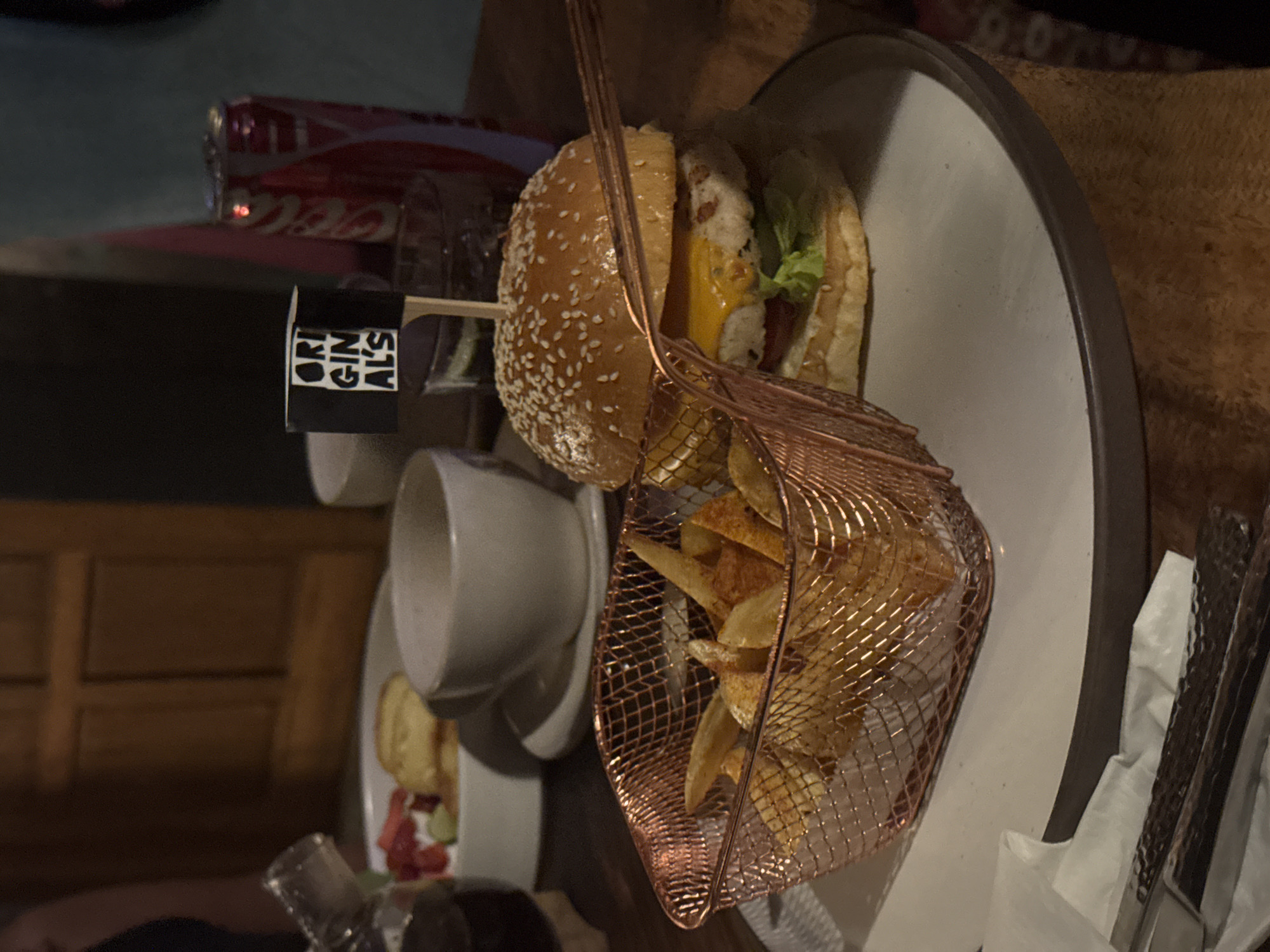 The image shows a plate with a burger and fries. The burger is on a sesame seed bun with lettuce, tomato, and cheese. The fries are in a copper-colored wire basket. There is a cup of coffee, and a bottle of Coca-Cola in the background. The plate is on a dark wooden table. The name of the restaurant is "Originals". The location is Unknown, Unknown.