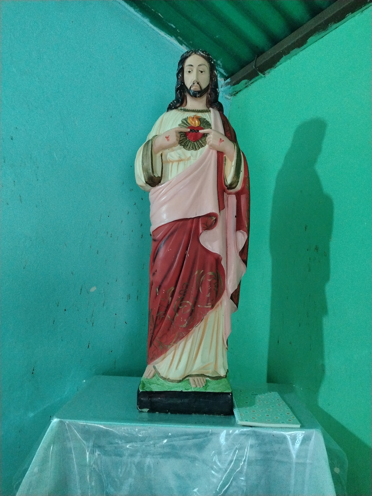 A full-length statue of Jesus Christ stands on a table covered with a clear plastic tablecloth in the corner of a room with light-green walls. Jesus is depicted with long, dark hair and a beard, wearing a cream-colored robe draped with a pink and red cloth. His hands are raised, pointing towards a stylized heart with flames emanating from it on his chest. A notebook lies on the table next to the statue's base. A tall, dark shadow of the statue is cast on the right wall. There is no indication of the statue being in Los Leoncitos, Venezuela.