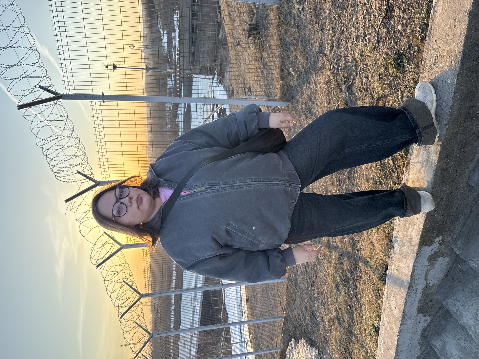 A young person with glasses stands facing the camera. They are wearing a gray jacket, a pink shirt, and dark jeans with gray cuffs. They also have white sneakers. A black bag is slung across their torso. Behind them is a chain-link fence topped with barbed wire. In the background, through the fence, is a field with patches of snow. The sky is visible in the distance and shows a sunset. The location is believed to be in Chaikovsky, Russia, based on visual features.