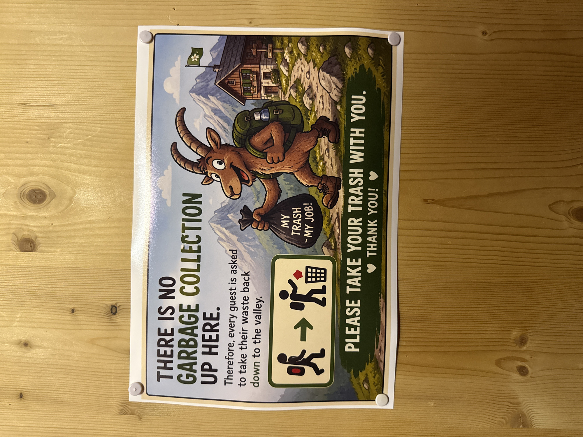 A sign is mounted on a wooden wall. The sign depicts a cartoon ibex carrying a backpack and a bag of trash. The ibex is walking on a mountain path towards a cabin. The text on the sign reads: "THERE IS NO GARBAGE COLLECTION UP HERE. Therefore, every guest is asked to take their waste back down to the valley. PLEASE TAKE YOUR TRASH WITH YOU. THANK YOU!" The sign is held up by four round, silver tacks. The wooden wall is made of vertical planks with visible wood grain and knots.