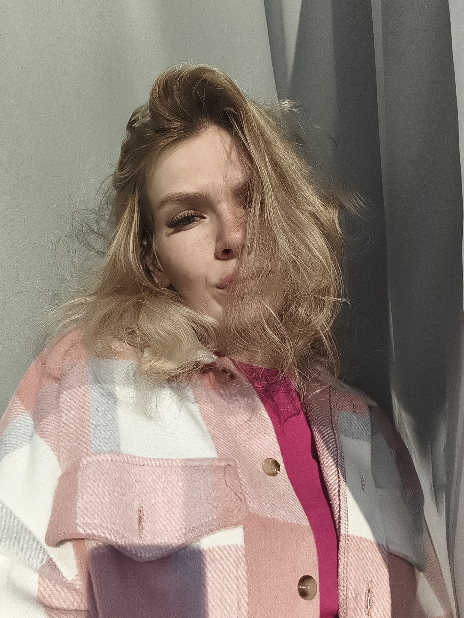 A close-up indoor portrait captures a young woman with long, wavy blonde hair, looking directly at the viewer. She has light skin and prominent eyelashes on her visible right eye. Her expression is playful, with her lips puckered into a "kissy face." Strong, direct natural light, likely from a window to her left, illuminates her face and hair, creating bright highlights and distinct shadows across her right cheek and the side of her body.
She is wearing a textured, thick plaid jacket in shades of pink, white, and light grey, over a vibrant hot pink top. Two brown-gold buttons are visible on the front of her jacket. The background consists of a plain light grey wall on the left and a darker grey curtain or fabric on the right. The intense, angled sunlight suggests it is either morning or late afternoon on a sunny day. The scene is set indoors, in Русский Вожой, Russia. No text is visible in the image.