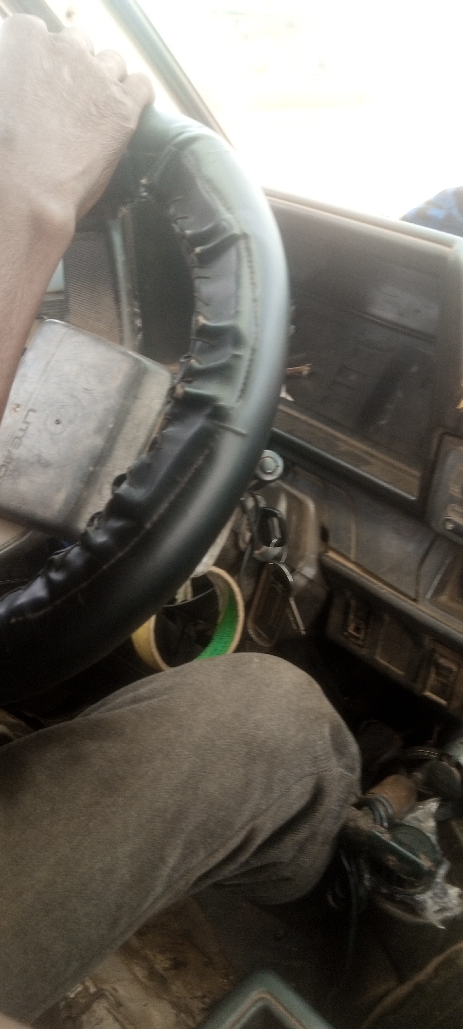 A person is driving a vehicle. The driver's left hand grips the steering wheel which has a worn black cover. A roll of tan tape with green markings is visible near the base of the steering wheel. Keys are in the ignition. The dashboard is old and dusty. The person is wearing gray pants. The location is Television, Nigeria, determined by metadata associated with the image.