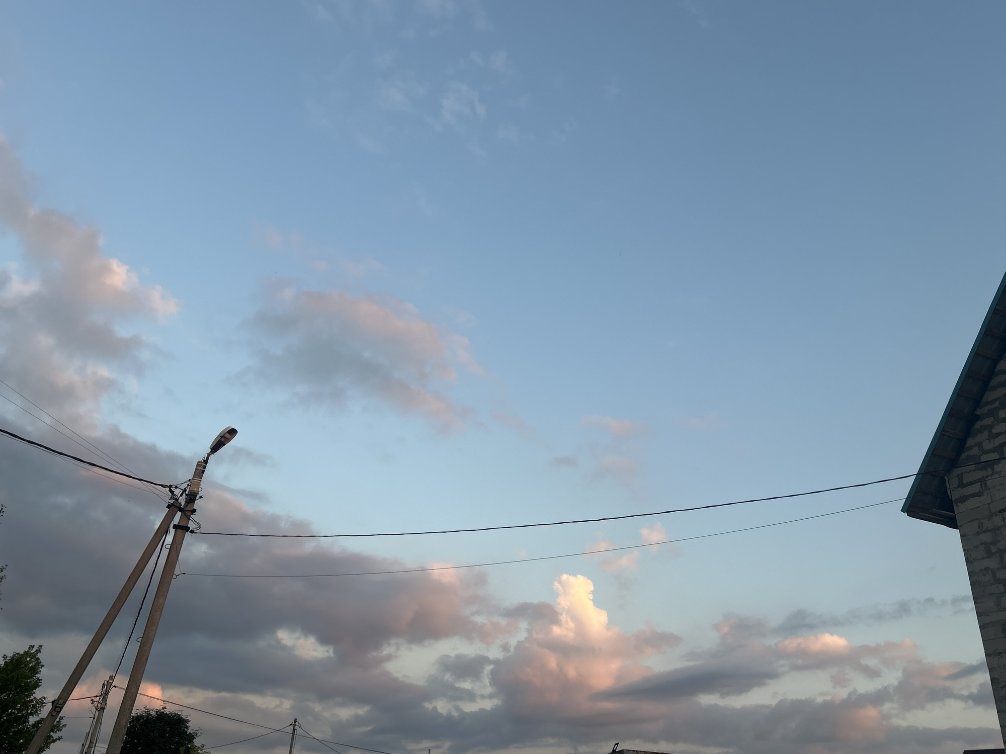 The image captures a twilight scene with a pale blue sky dotted with wispy clouds tinged with pink and gray. In the foreground, a tall, weathered utility pole stands at an angle, supporting electrical wires and a streetlamp. The pole is made of concrete and has a visible support structure. To the right, the corner of a building constructed of light-colored bricks and topped with a dark sloped roof is visible. The overall impression is of a residential or semi-rural area in Нижние Муллы, Russia, during the early evening. No people are present in the frame.