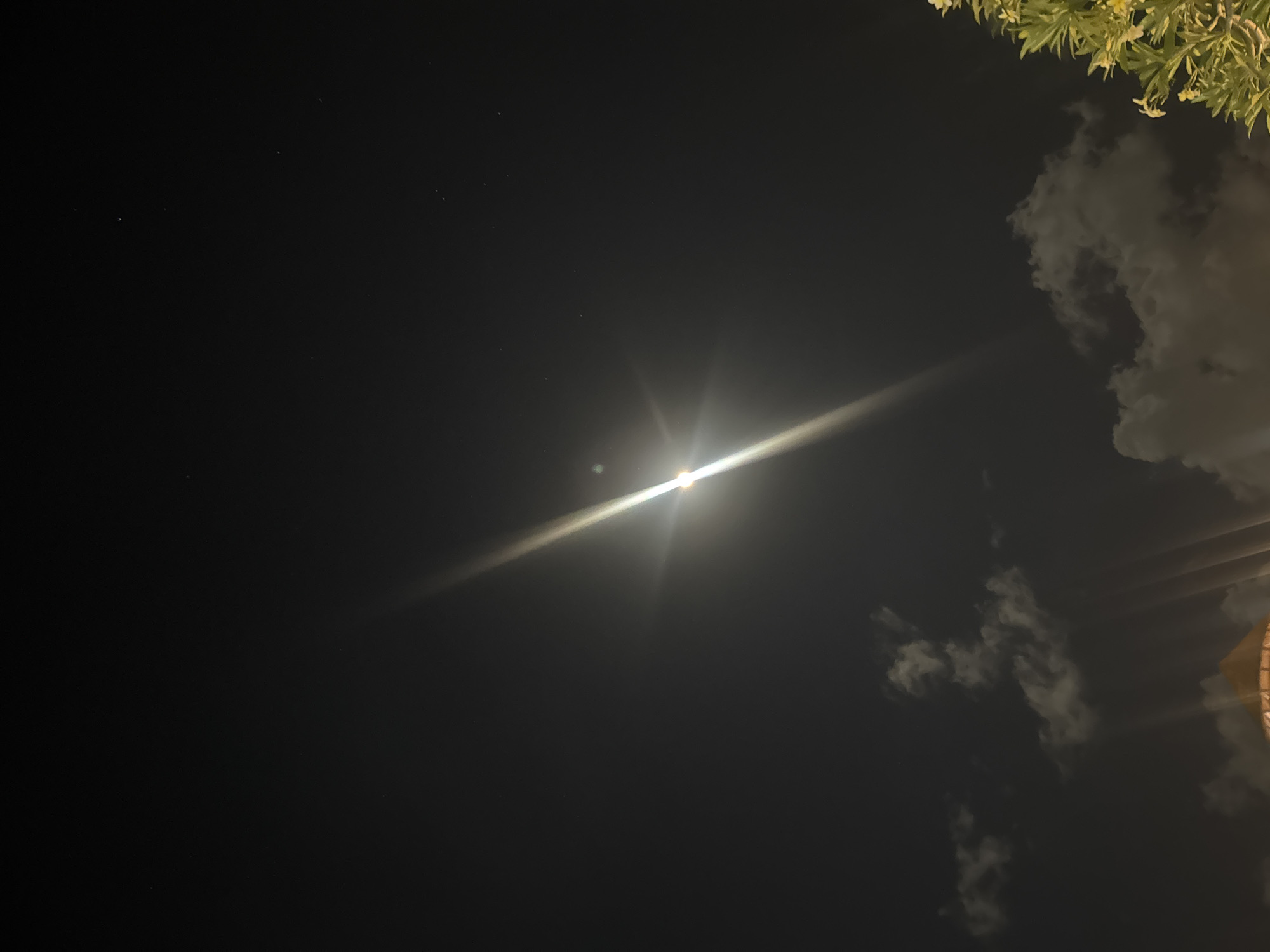 A nighttime exterior shot taken from a low angle looking upwards at the sky. The scene captures a moonlit night with a bright, lens-flared moon positioned in the upper center of the frame. Scattered clouds are visible across the lower half of the sky, and a few faint stars are present in the dark expanse above. At the bottom of the image, the top of a building with a peaked roof and lights emanating from it can be seen. To the right, the branches and leaves of a tree with small white flowers are illuminated by artificial light. The location is Kuta, Indonesia.