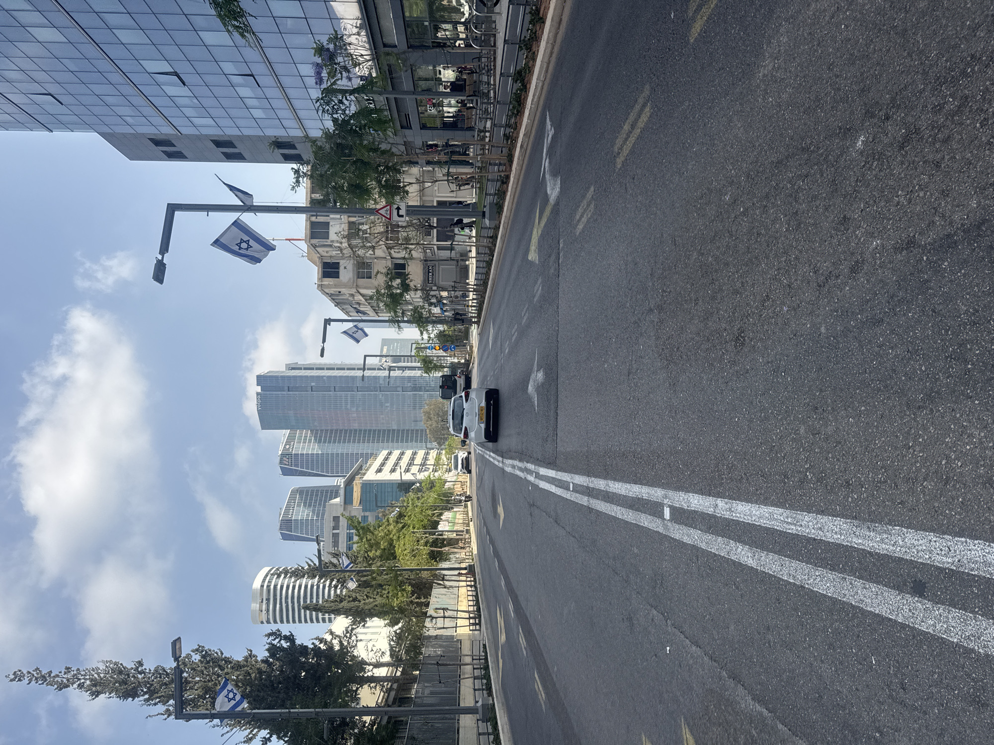 The image shows a street in Tel Aviv, Israel. The road is asphalt with double white lines marking the center. There are buildings of varying heights on either side of the street, including modern skyscrapers and older, less imposing structures. Israeli flags are affixed to lampposts along the side of the road. There are trees and vegetation interspersed between the buildings. A white car is visible on the road, moving away from the camera. The sky is blue with scattered clouds.