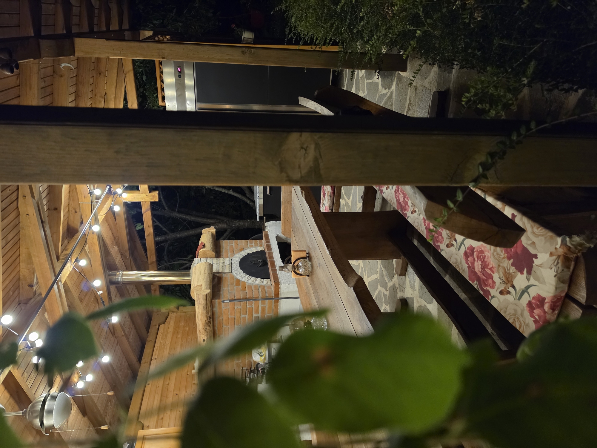 The image shows an outdoor dining area at night, likely a patio or backyard, with wooden structures and string lights. In the center, a brick pizza oven with a round opening is visible, suggesting it's ready for use or has recently been used. A metal flue pipe extends from the oven. A wooden table with a floral-patterned tablecloth is set up, with chairs that appear to have cushions. Stone flooring is visible in the background. Lush green foliage is in the foreground, partially obscuring the view. The scene is illuminated by warm string lights, creating a cozy ambiance. The time of day is clearly night, and the weather appears to be clear. There are no people visible in the image. The overall impression is one of a relaxed, inviting outdoor space designed for gatherings and dining.