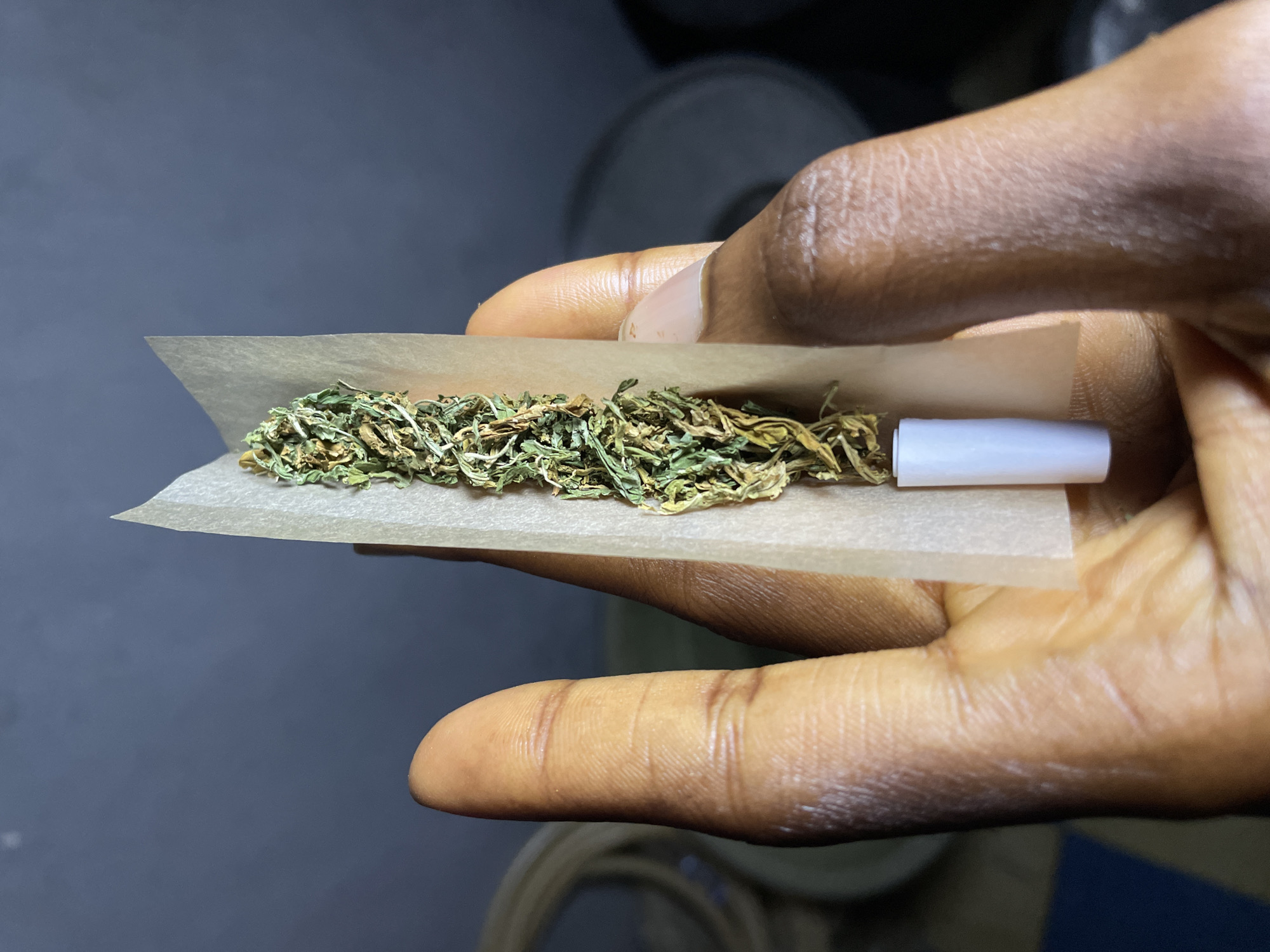 In Nkpor, Nigeria, a close-up shot captures a person's hands holding a marijuana cigarette being rolled. The fingers, with dark skin and visible wrinkles, are carefully manipulating a thin rolling paper filled with dried cannabis. A white filter tip is attached to one end of the paper. The background is dimly lit and out of focus, with hints of circular objects and a dark wall.