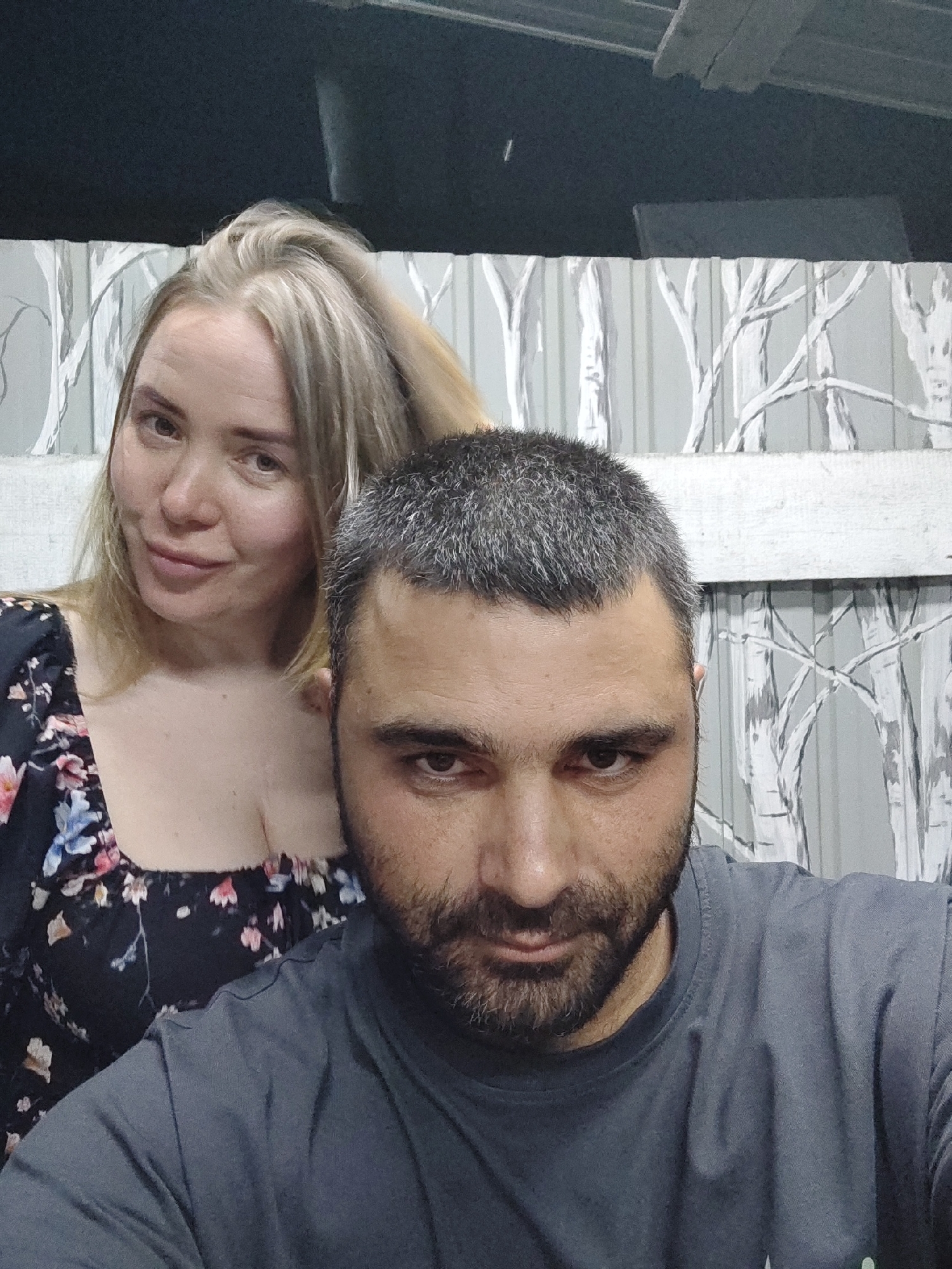 A medium shot shows a couple posing for a selfie. The man is in the foreground, his face close to the camera, and a woman is behind him, her head tilted slightly. The background consists of a fence with a stylized white tree pattern. They are outdoors, possibly in the city of Shimanovsk, Russia, given the context. The man has short, graying hair and a beard, wearing a gray t-shirt. The woman has blonde hair and is wearing a dark floral dress. The lighting is dim, suggesting it is evening or night.
