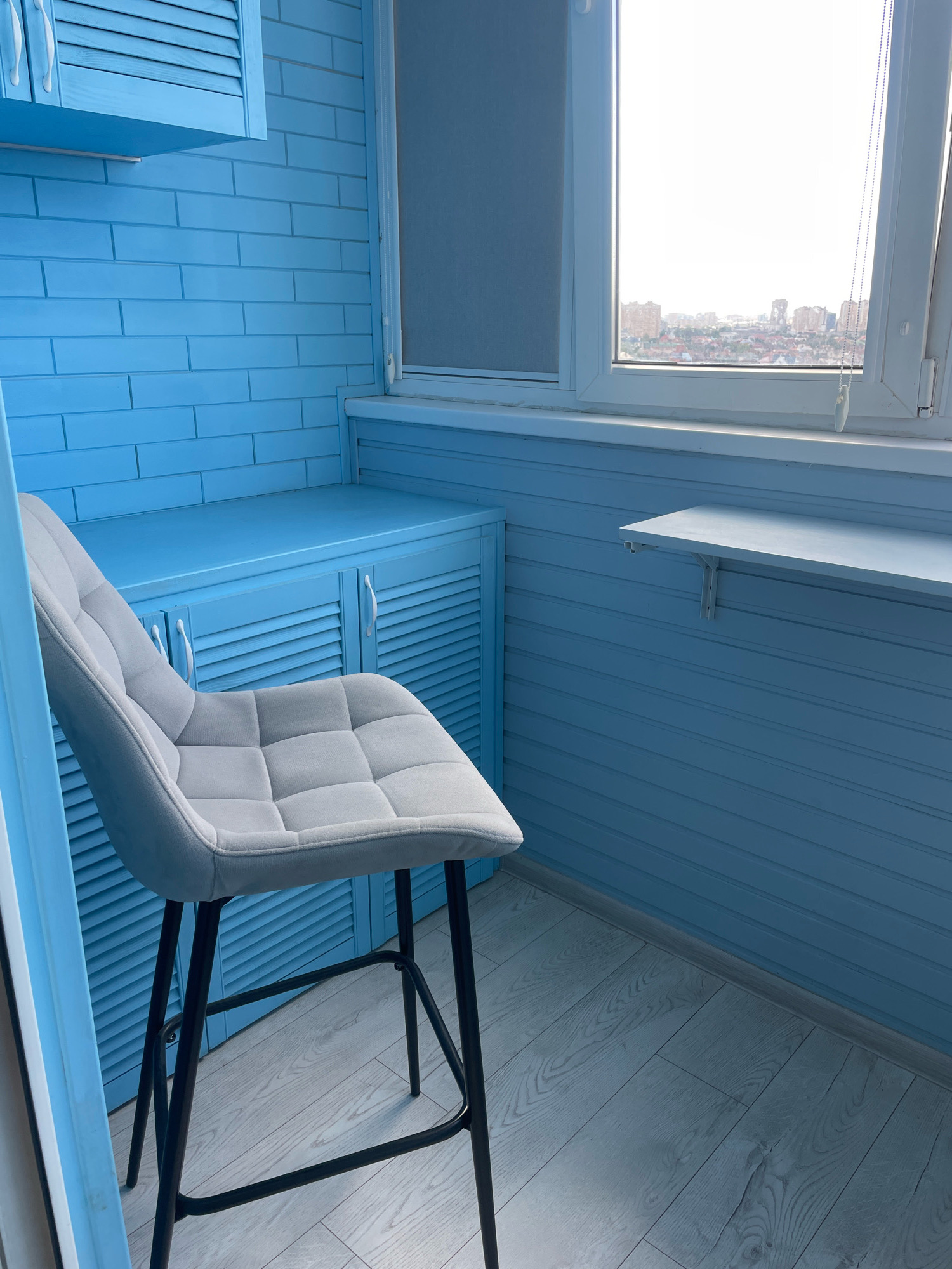 The image depicts the interior of a balcony, likely in Krasnodar, Russia, given the context. The balcony walls and cabinets are painted a bright, uniform blue. The walls appear to be clad in horizontal planks, and a section also features a brick pattern. To the left, a blue cabinet with louvered doors and white handles is visible. In the foreground, a light gray upholstered bar stool with a tufted seat and black metal legs stands on a light-colored wooden laminate floor. A fold-down table, also light blue, is mounted on the wall above the cabinets. Through the window, a cityscape with buildings and a bright sky can be seen. The scene is brightly lit, suggesting daytime. No people are present in the image.