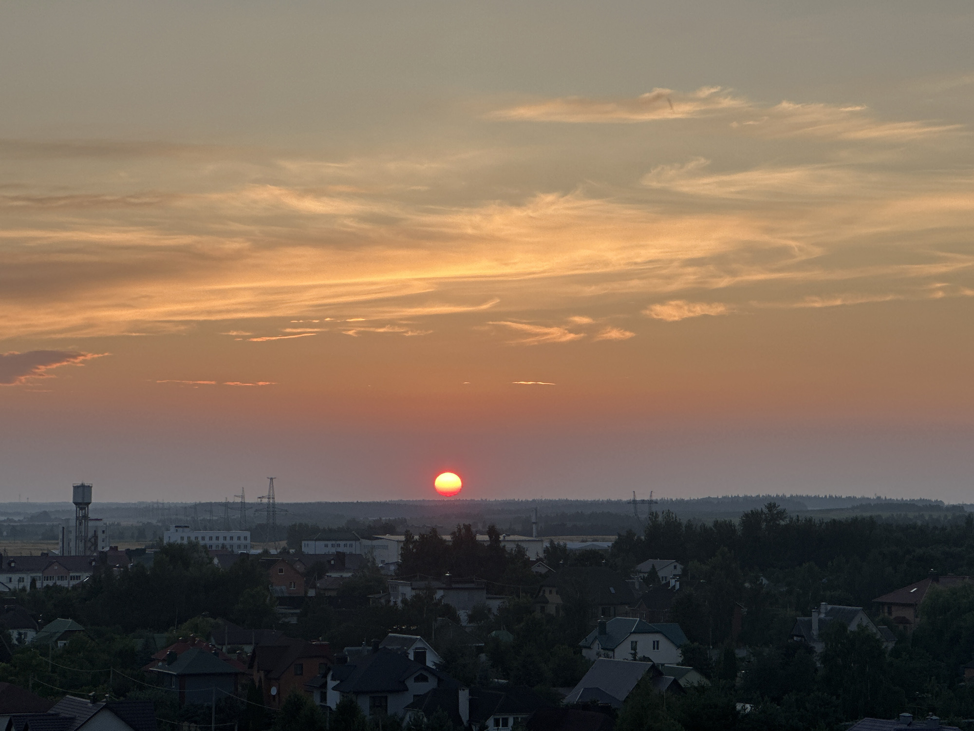 The image captures a sunset over the city of Shchomyslitsky Selsoviet, Belarus. The sun, a bright orange orb, is positioned near the center of the frame, low on the horizon. Wispy clouds, tinged with orange and yellow hues from the setting sun, stretch across the sky. Below the sky, a panoramic view of the city unfolds. Rooftops of houses, interspersed with dense green trees, dominate the foreground and middle ground. In the distance, industrial structures, including a water tower and power lines supported by tall pylons, are visible, silhouetted against the twilight sky. The overall atmosphere is serene, with the warm glow of the sunset casting a peaceful ambiance over the urban and natural landscape.