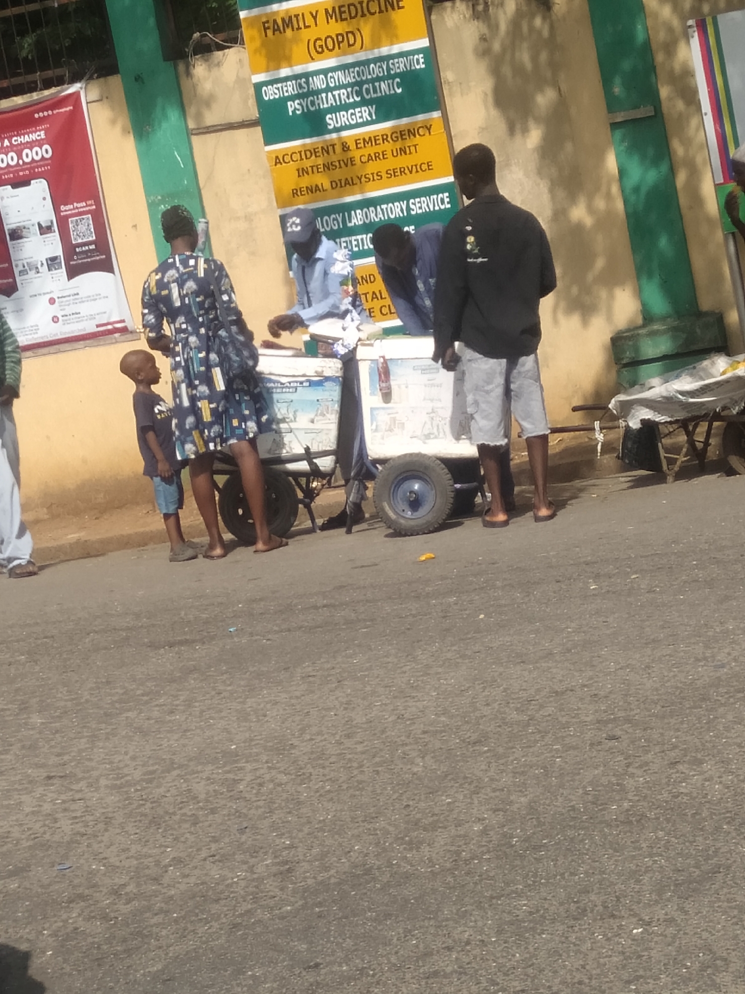 An outdoor scene captured during the daytime shows several individuals gathered around a mobile cart on a paved road. In the foreground, a woman in a dark patterned dress stands with her back to the camera, next to a small boy in a dark t-shirt and light shorts, who is looking towards the cart. Behind a white mobile cart with wheels, two men are present. One, wearing a light blue shirt and a cap, leans over the cart, while another in a black long-sleeved shirt and light shorts stands upright. A partial view of another person is visible on the far left. To the right, an additional older cart or trolley with a white covering is visible.
The background features a large yellow and green sign that details various medical services, indicating the location is likely outside a hospital or clinic. The sign reads: "FAMILY MEDICINE (GOPD)", "OBSTERICS AND GYNAECOLOGY SERVICE", "PSYCHIATRIC CLINIC", "SURGERY", "ACCIDENT & EMERGENCY", "INTENSIVE CARE UNIT", "RENAL DIALYSIS SERVICE", "LOGY LABORATORY SERVICE", and "JETETIC". Adjacent to this, on a red banner, text such as "0000", "A CHANCE", "00,000", "Gate Pass", and "SCAN ME" is visible. The white cart also has "AILABLE HERE" printed on its side. Bright sunlight casts clear shadows on the ground, suggesting a sunny day.