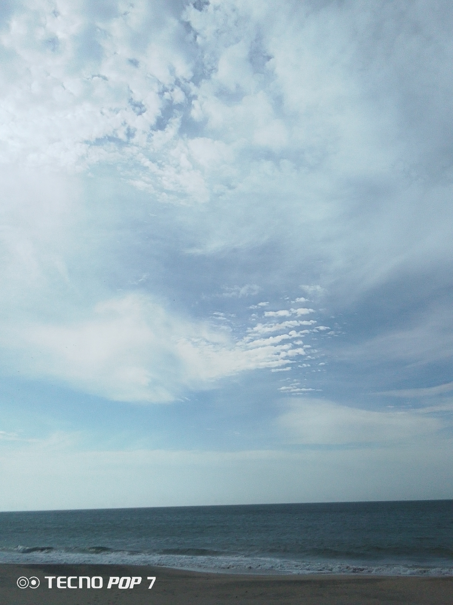 The image depicts a coastal scene in Pedro González, Venezuela. The sky is predominantly visible, filled with light blue and white clouds arranged in varied patterns. The ocean occupies a smaller portion of the frame, with a dark blue hue and small waves breaking near the shoreline. A narrow strip of sand is visible at the bottom. "Tecno Pop 7" is printed at the lower left corner of the image. No people are visible.
