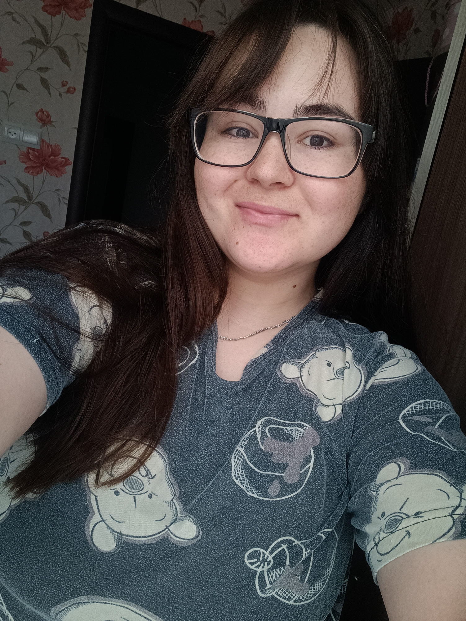 A young adult woman with long, dark brown hair and glasses is captured in a selfie-style shot, smiling gently at the camera. She wears a dark blue t-shirt or pajama top patterned with light-colored, stylized cartoon characters, including what appear to be Winnie the Pooh. A thin, silver-colored chain necklace is visible around her neck.
The setting is indoors, likely a residence in Noviy, Russia. Behind her, on the left, is a wall adorned with light-colored wallpaper featuring a repeating pattern of large, red/orange flowers with green leaves. A white electrical outlet with two sockets is visible on this wall. To her right and behind her, a dark doorframe or piece of wooden furniture is visible. The lighting is soft and even, suggesting indoor light or diffused daylight. The woman has noticeable freckles across her nose and cheeks. Her expression conveys a sense of calm and friendliness. The cartoon characters on her top are depicted in a somewhat distressed or hand-drawn style, some appearing within circular outlines.
