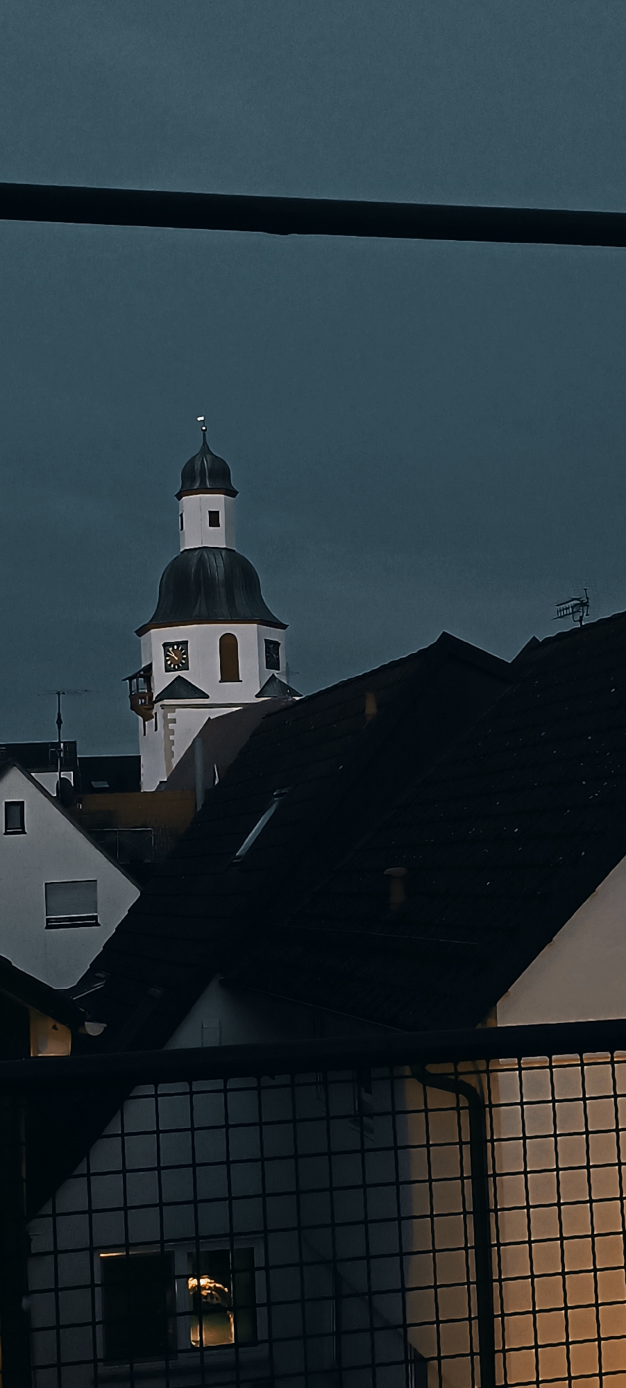 The image shows a dark, twilight scene with a view of the tower of the Church of St. Michael in Winnenden, Germany. The tower has a white base with a dark, domed roof and a spire. There are clocks visible on the tower. In the foreground, dark rooftops of houses are visible, some with skylights. The sky is a deep, dark blue, suggesting dusk or early evening. The scene is partially obscured by a black metal fence or railing in the very foreground. There are no people visible in the image. The overall impression is one of a quiet, possibly overcast evening in a town.