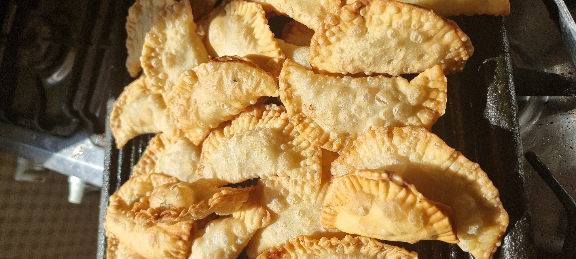 A close-up, top-down shot displays a pile of golden-brown, fried empanadas arranged on a dark, textured surface. The empanadas, varying in size and shape, are golden and appear to be made of a flaky dough, with visible crimped edges. Some empanadas exhibit small bubbles and blisters on their surface, indicating they have been freshly fried. Sunlight illuminates the empanadas from the upper left, casting subtle shadows and highlighting their crisp texture. The background is out of focus, showing hints of metallic and textured elements, possibly a stovetop or grill. The overall scene is focused on the appetizing appearance of the fried pastries. There is no indication of the location being Aktobe, Kazakhstan.