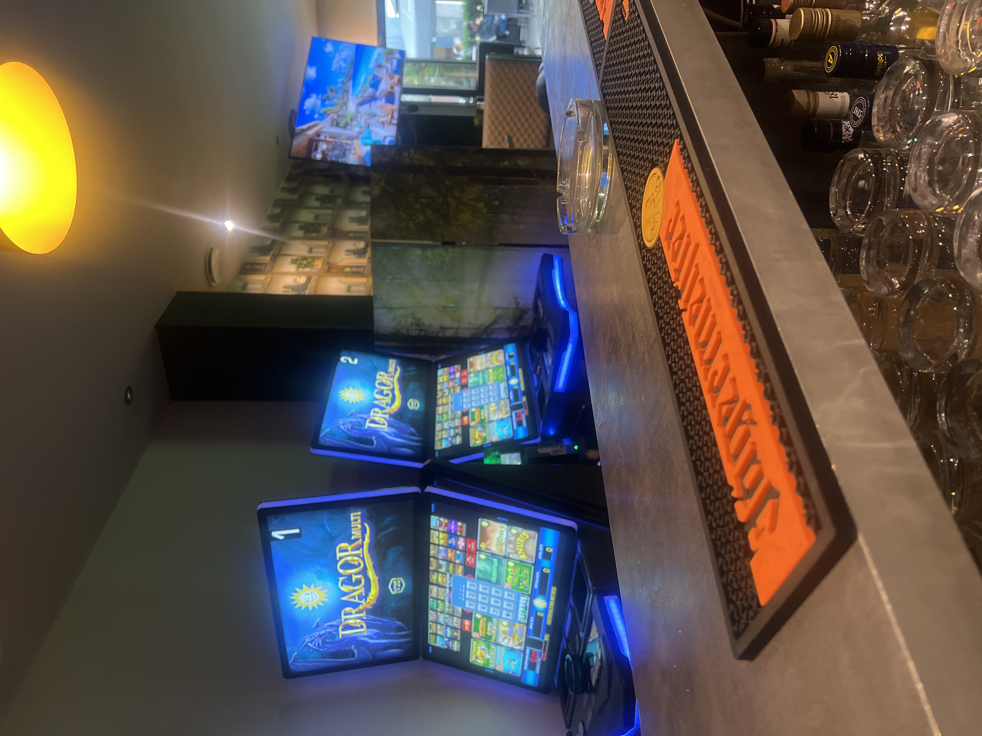 The image depicts the interior of a bar or casino in Ludwigshafen am Rhein, Germany. Two "DRAGOR MULTI" slot machines are prominently featured, illuminated by blue neon lights. The machines display various game interfaces. In the foreground, a bar counter is visible, with an ashtray and a "Jägermeister" branded bar mat. Behind the bar, shelves stocked with bottles and glassware can be seen. A television screen displaying a tropical scene is mounted on the wall in the background. The lighting is dim, with a bright yellow light fixture visible in the upper part of the frame. The environment appears to be a commercial establishment, likely catering to gaming and entertainment.
