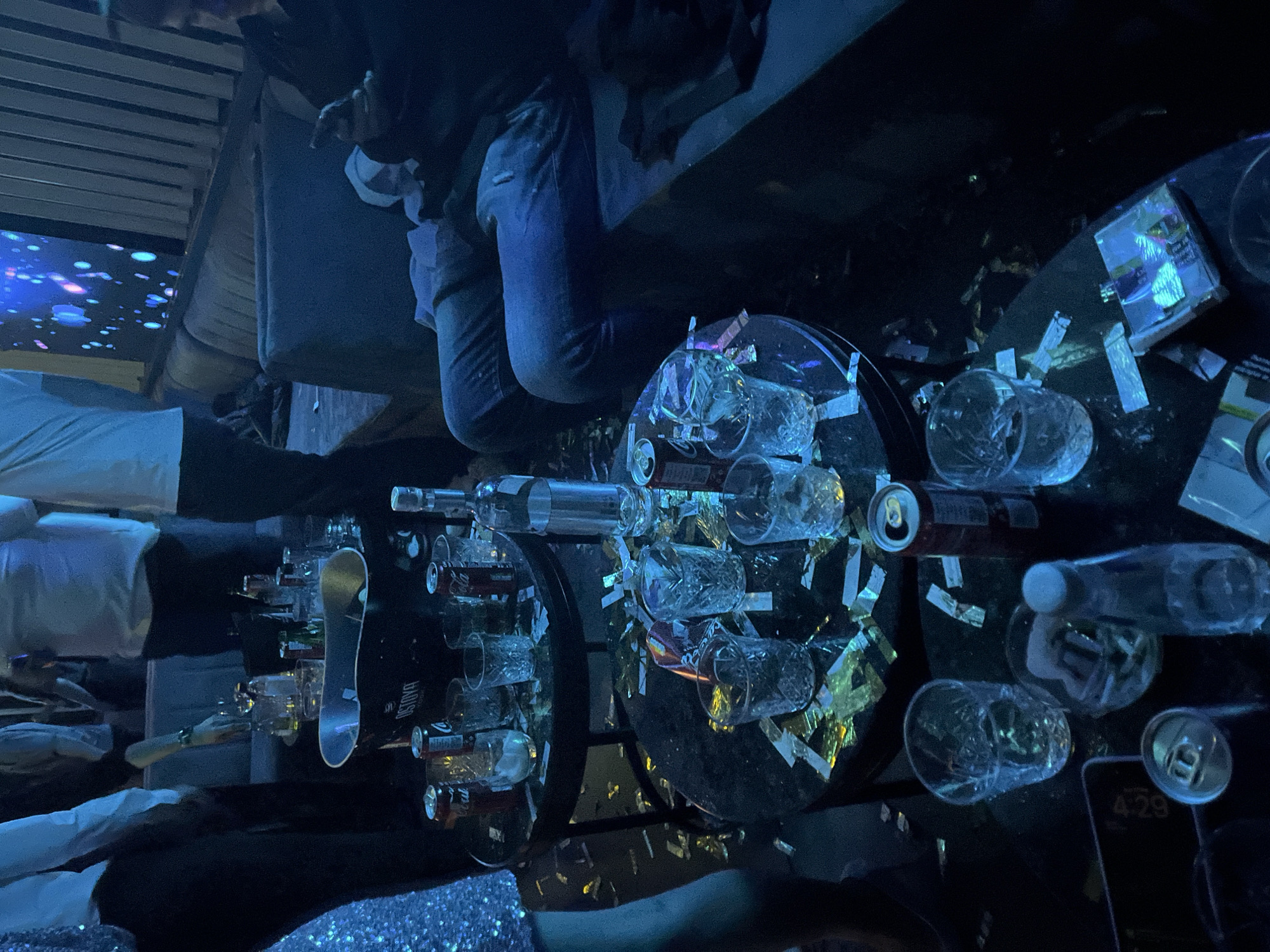 The image depicts a scene likely inside a nightclub or bar in Plovdiv, Bulgaria. Tables are cluttered with empty bottles and cans of beverages. There are glasses and scattered confetti. A person in jeans is sitting on a nearby surface. A smartphone shows the time 4:29. There's a screen with a blue, swirling pattern in the background.
