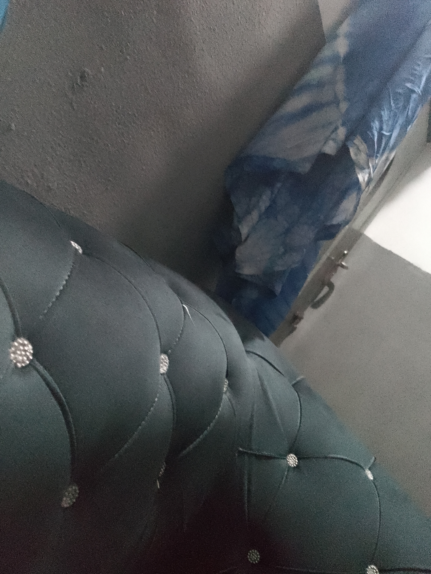 The image displays a close-up, angled view of a dark teal upholstered couch with tufted detailing and crystal embellishments. The couch occupies the foreground and mid-ground of the image. In the background, a textured grey wall is visible, and to the right, a rolled-up blue object, possibly bedding or fabric, is leaning against the wall, partially wrapped in plastic. The setting appears to be indoors, likely a room. There is no indication of Jauro Yinu, Nigeria, or any specific location.
