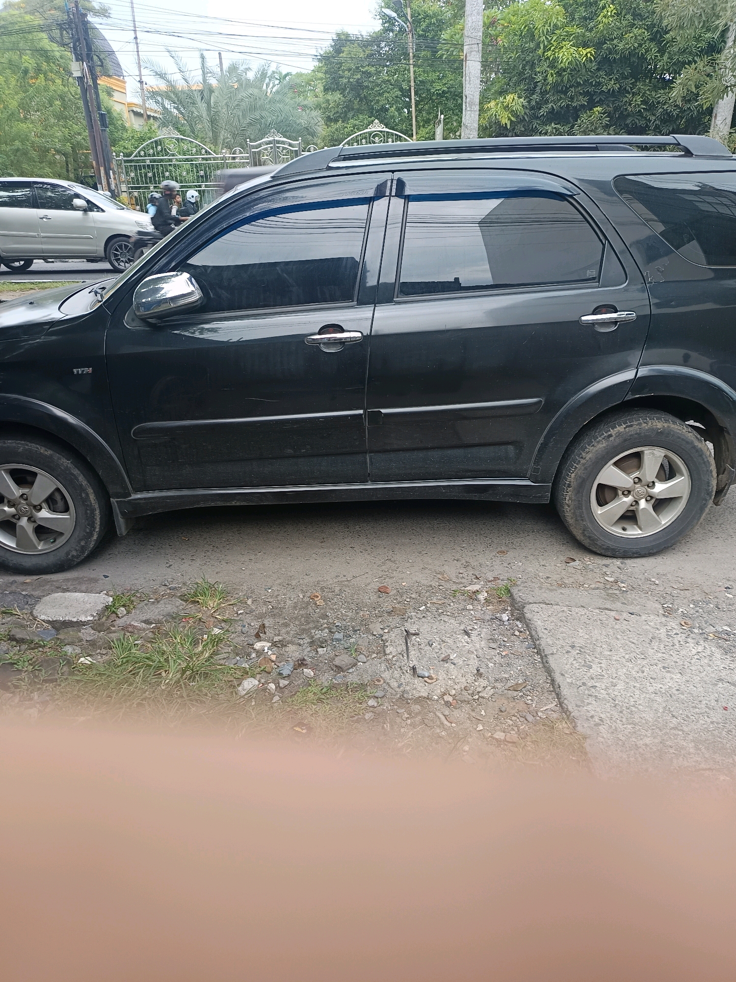 The image captures a side view of a dark-colored SUV parked on a dirt and concrete roadside in Medan, Indonesia. The vehicle, which appears to be older, has a roof rack and tinted windows. In the background, a silver car is visible, and a motorcycle with two people wearing helmets is in motion on the road. The environment includes trees, power lines, and a decorative metal fence. The lower portion of the image is blurred, obscuring what might be a person's hand or a similar obstruction.