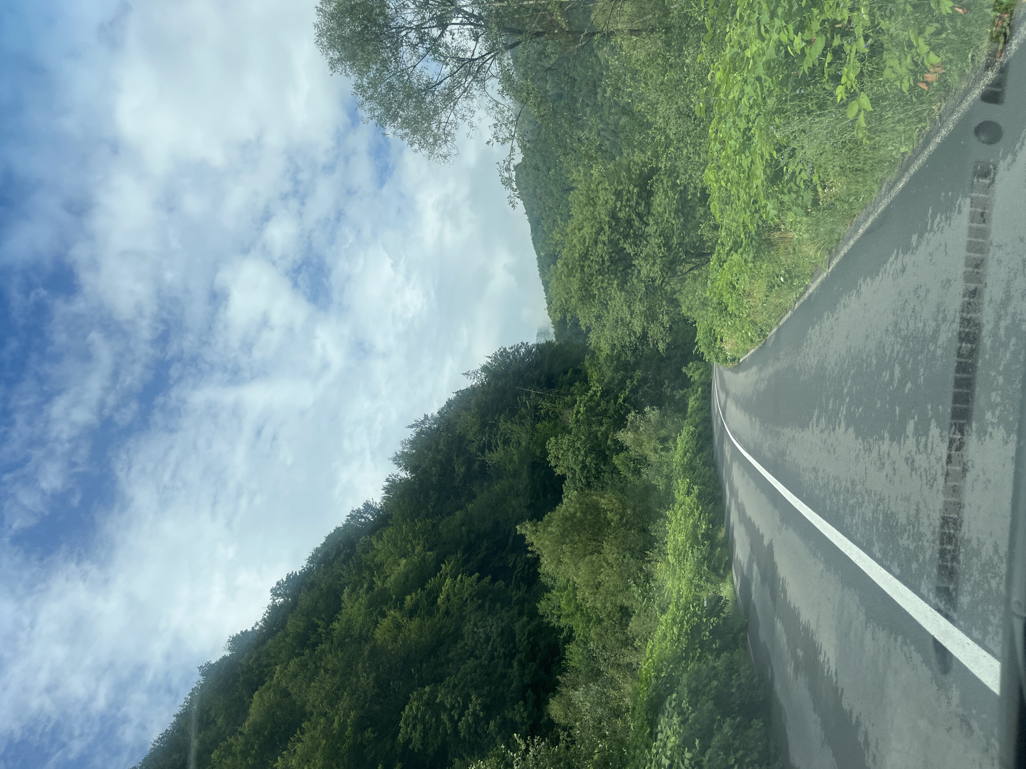 The image depicts a scenic view of a winding asphalt road with a white dividing line, passing through a lush, green mountainous landscape. The road appears wet, suggesting recent rain. Dense forests cover the slopes of the mountains on both sides of the road. The sky above is partly cloudy with scattered white clouds against a blue background. The perspective suggests the photo was taken from within a vehicle, looking forward along the road. The location is identified as Kostylivka, Ukraine. There are no people visible in the image.