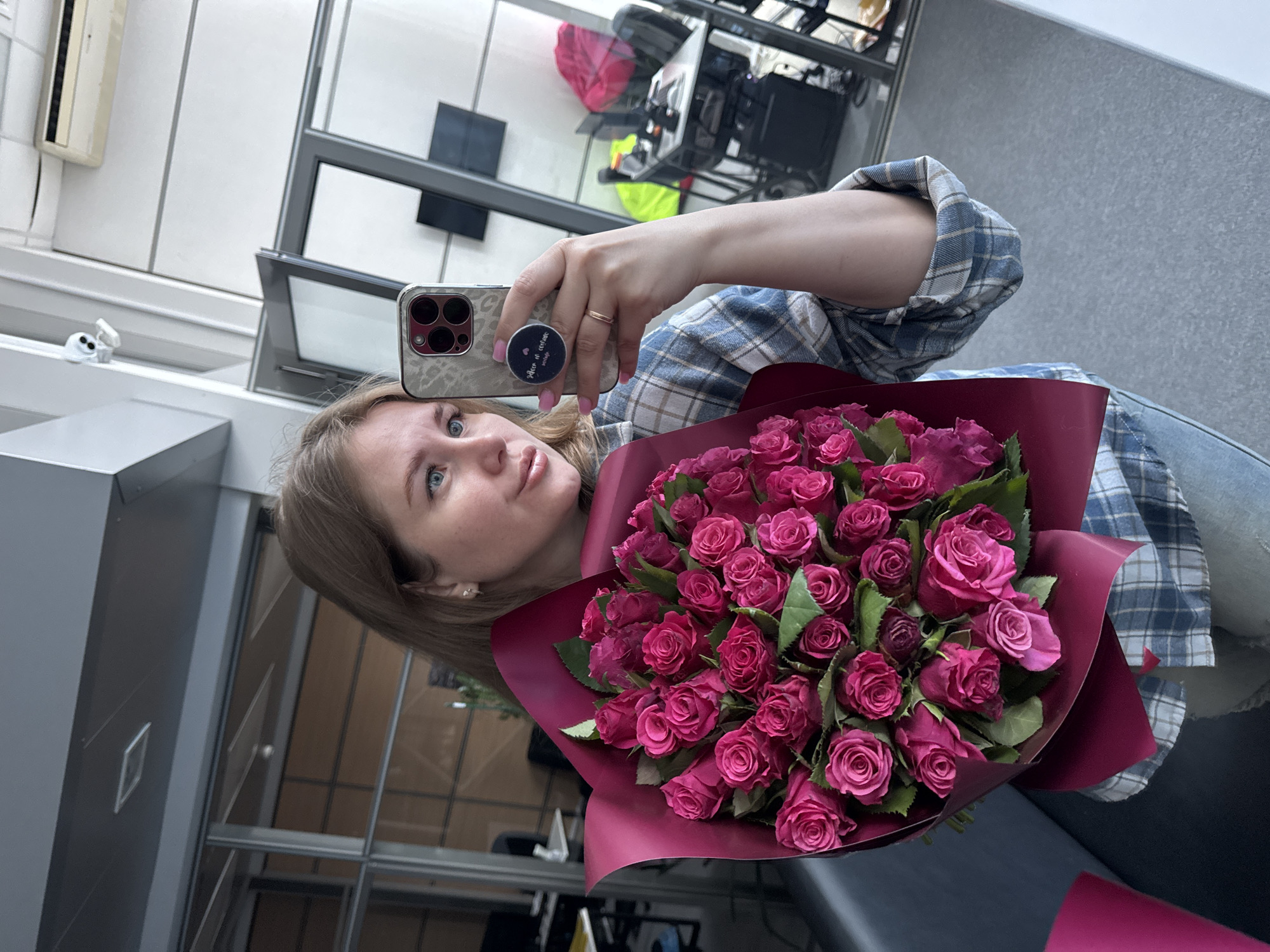 A blonde woman with blue eyes is taking a selfie in a modern office environment. She is holding a large bouquet of pink roses. She is wearing a blue and white checkered shirt and blue jeans. In the background, there are office desks, chairs, computers, and windows. The location is identified as an office in Perm, Russia.