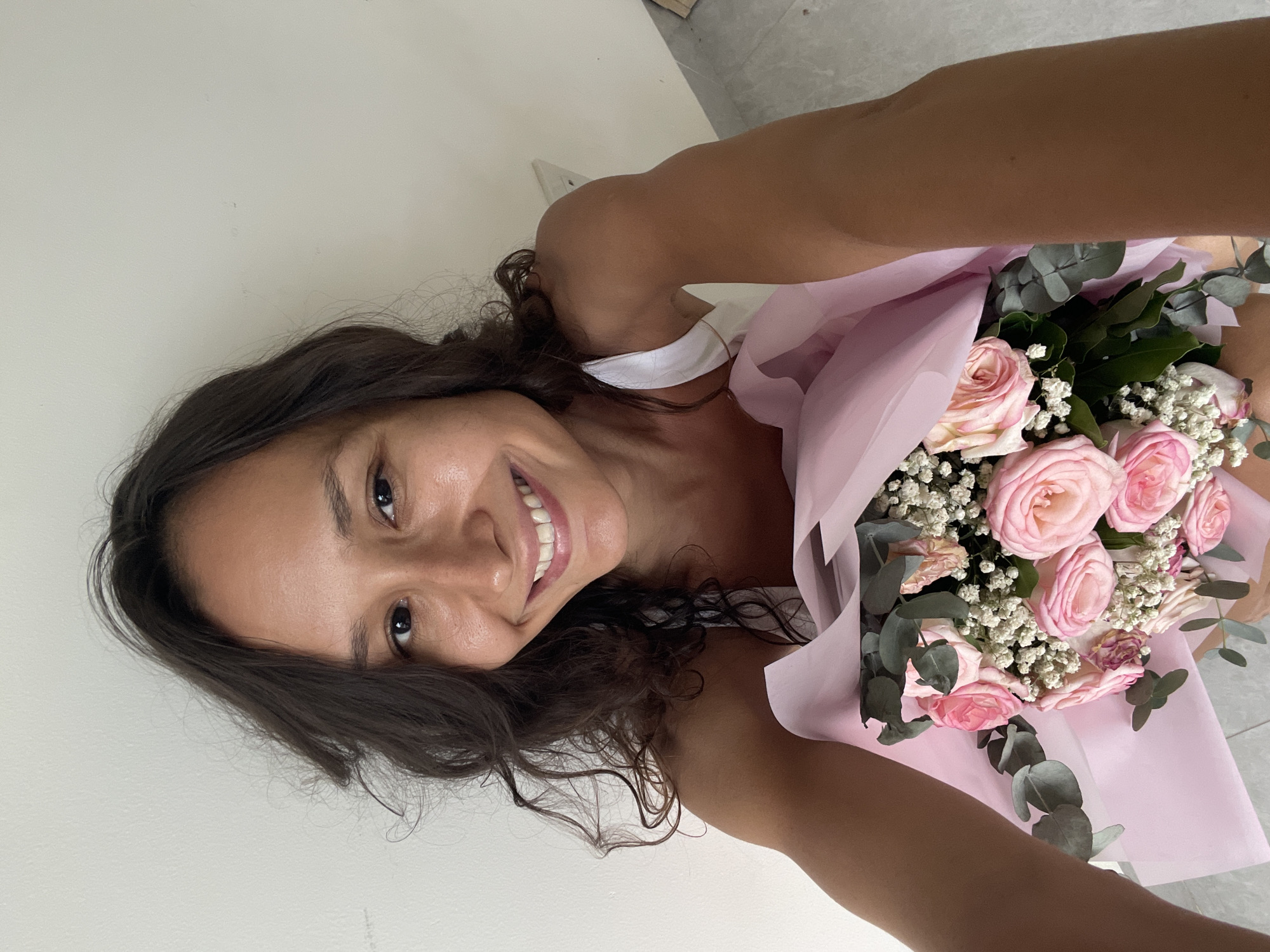 A woman, with dark, curly hair and fair skin, smiles broadly at the camera while holding a bouquet of pink roses and eucalyptus. She is wearing a white tank top. The background is a plain, off-white wall. The lower portion of the image shows the bouquet, which is wrapped in pink paper. The scene appears to be indoors, possibly a hotel room or apartment, with a hint of a tiled floor visible at the bottom right. The lighting is natural, suggesting daylight. There is no discernible connection to the city of Mũi Né, Vietnam.