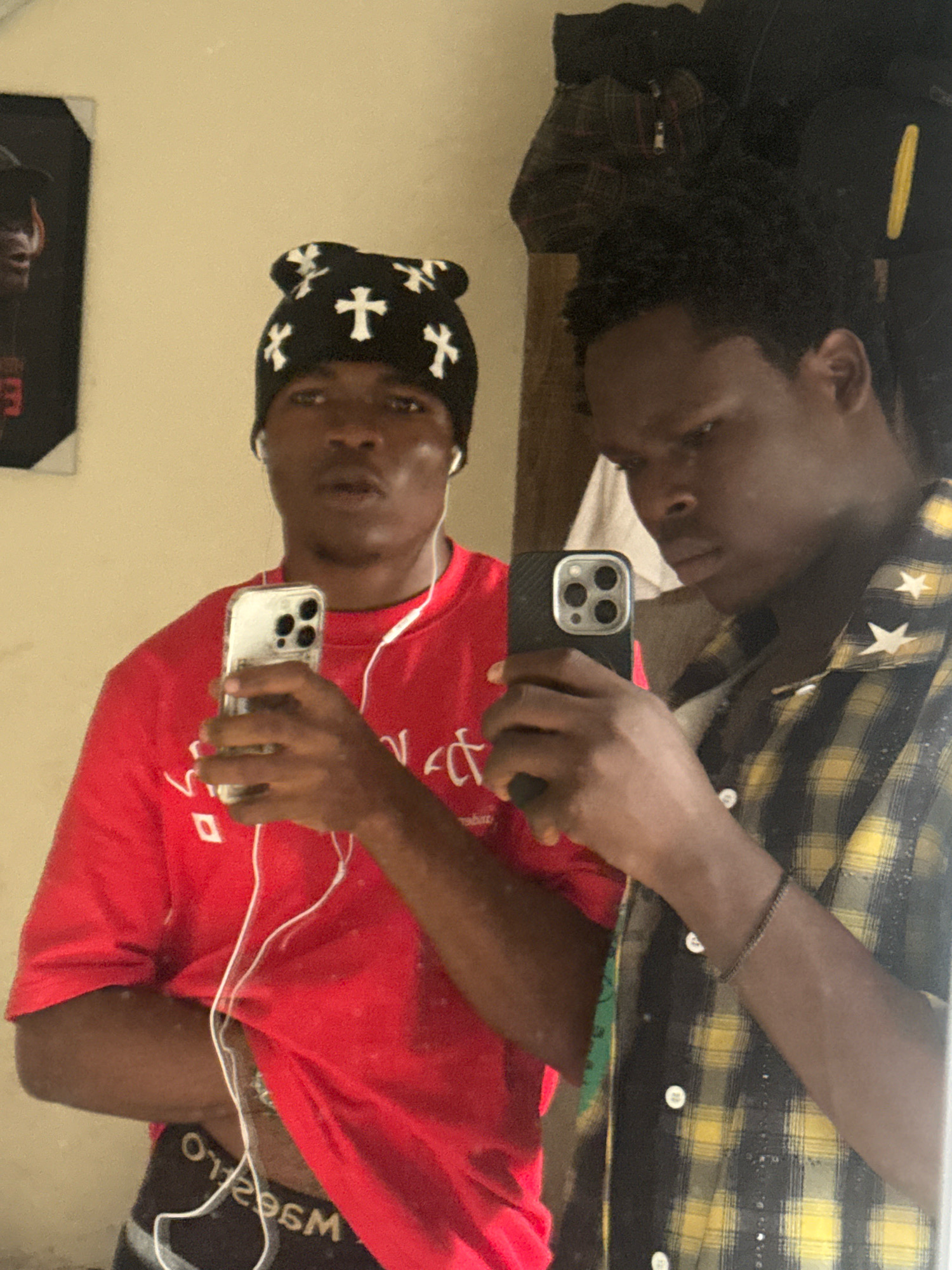 Two young men are taking a mirror selfie in what appears to be a room. The man on the left is wearing a black beanie with white crosses, a red t-shirt, and appears to be holding a smartphone. The man on the right is wearing a yellow and black plaid shirt and is also holding a smartphone. The background is simple, with a plain wall and some items hanging on it. The lighting suggests an indoor setting. The image captures a casual moment between the two individuals. There is no indication of the specific location within the city of Ihiagwa, Nigeria.