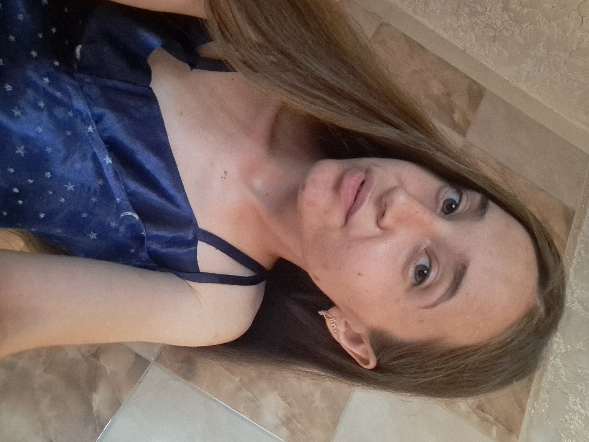 A young woman with long, brown hair is captured in a selfie, her gaze directed towards the camera. She is wearing a dark blue slip dress adorned with a pattern of white stars. Her right ear sports a silver earring. The background features a tiled wall with a marbled pattern, suggesting an indoor setting. The lighting is bright, illuminating her face and shoulders. The image was taken in the city of Vypasne, Ukraine.