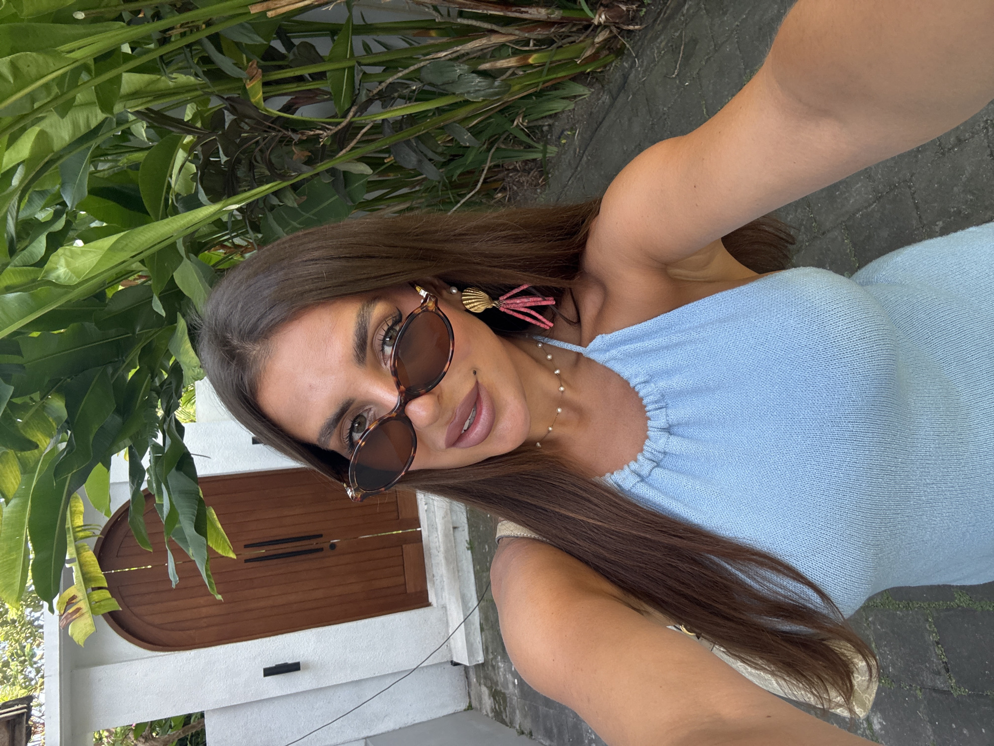 A woman is seen in a selfie outdoors in North Kuta, Indonesia. She has long, brown hair, sunglasses, and a light blue knit top. Her accessories include a shell earring with pink tassels, and a pearl necklace. Behind her is a wooden double door in a white wall, and various tropical plants. The ground appears to be paved.