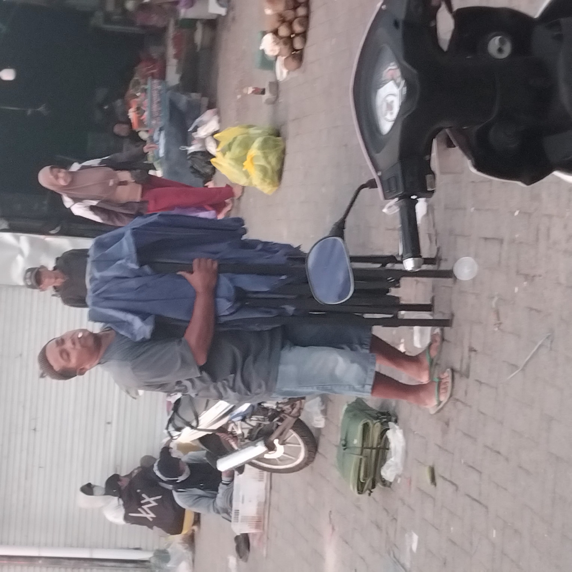 The image depicts a street scene in Banjarbaru, Indonesia, likely in a marketplace area. A man in a grey t-shirt and denim shorts is holding a folded blue tarp or tent. He has a cigarette in his mouth. To his left, a motorcycle is parked. In the background, several people are visible. One person is wearing a dark hoodie and a cap. Another person, a woman wearing a hijab and red pants, is standing near a table with goods, possibly a vendor. There are stacks of what appear to be coconuts or similar round produce on the right side of the image. The ground is paved with square bricks. The overall atmosphere suggests an outdoor commercial area.