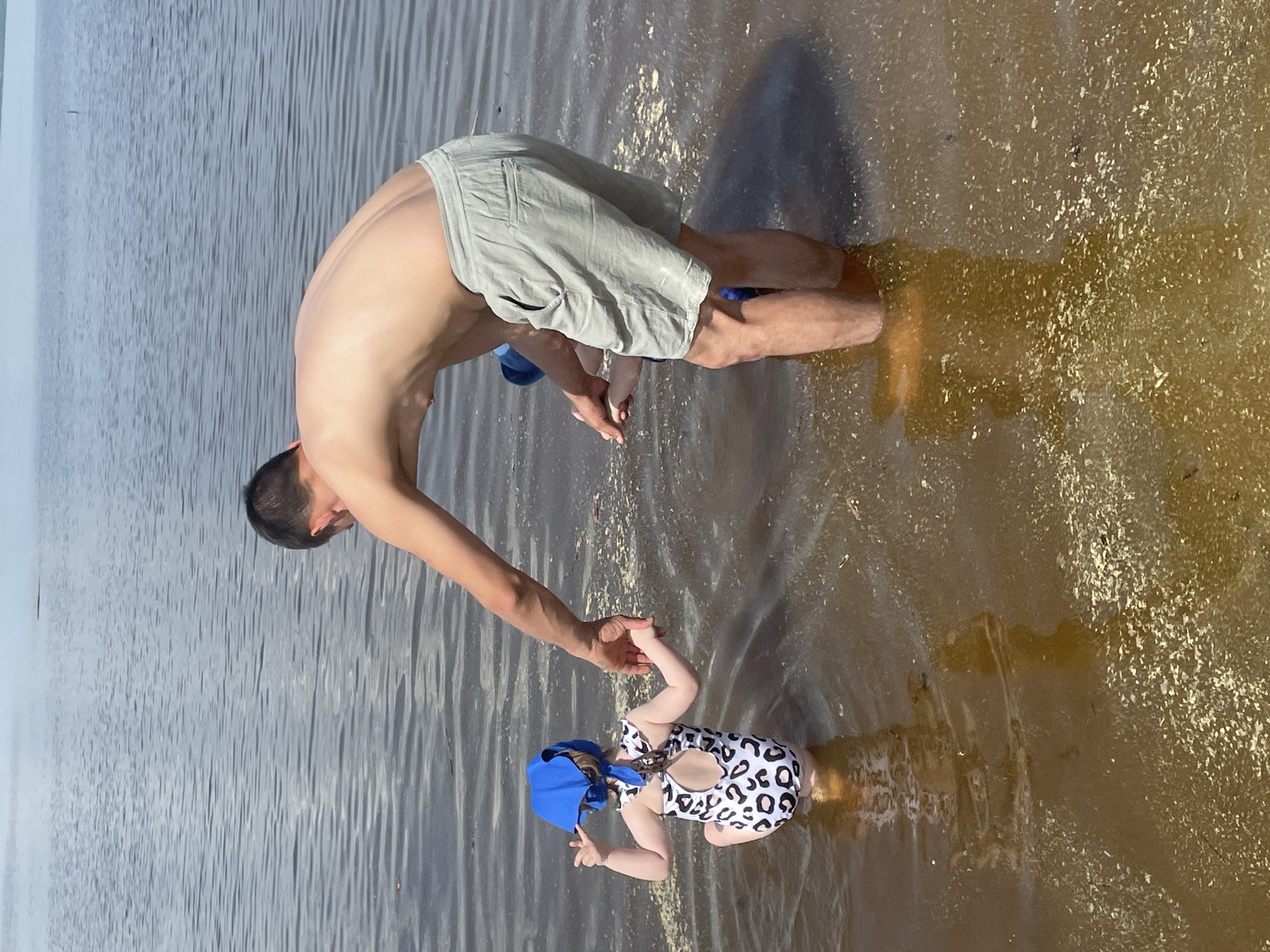 A shirtless, light-skinned man wearing light-colored shorts is wading in a body of water, bent over and holding hands with a young child wearing a blue cap and a white and black spotted swimsuit. The water's color transitions from grayish on the left to a brownish-gold on the right. Ripples are visible in the water.
The precise location in the city of Perm, Russia, is not discernible from the visual information provided.