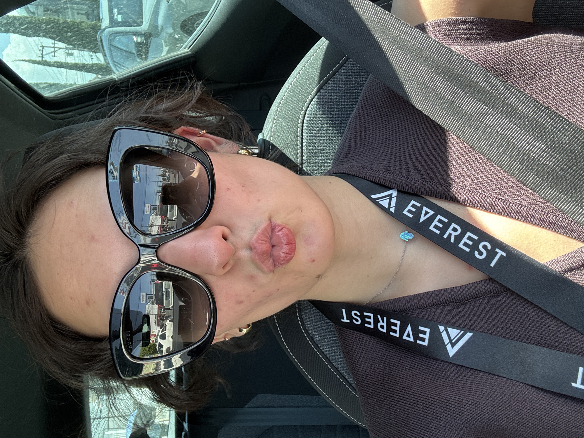 A female individual is depicted in a close-up selfie, seated within a vehicle. She has short, dark hair and wears black, cat-eye style "Gucci" sunglasses, obscuring her eyes. Her lips are pursed in a "kiss" gesture, and her visible facial skin exhibits minor blemishes. She wears gold hoop earrings in both ears, a dark brown ribbed top, and a black lanyard with "EVEREST" in white capital letters and a white triangular logo. A thin silver chain necklace with a small, light blue hamsa-shaped pendant is visible. A black seatbelt crosses her chest and lap, secured over a gray fabric car seat featuring white stitching.
The external environment, reflected in the sunglasses and visible through the vehicle's side window, presents an urban street scene. The left sunglass lens reflects a road with a white sedan, other vehicles, and a utility pole with overhead wires. Through the passenger side window, tall, slender trees, another utility pole with numerous wires, a light-colored SUV, and a blue sky with scattered clouds are visible. This scene is located in the city of Puebla, Mexico.