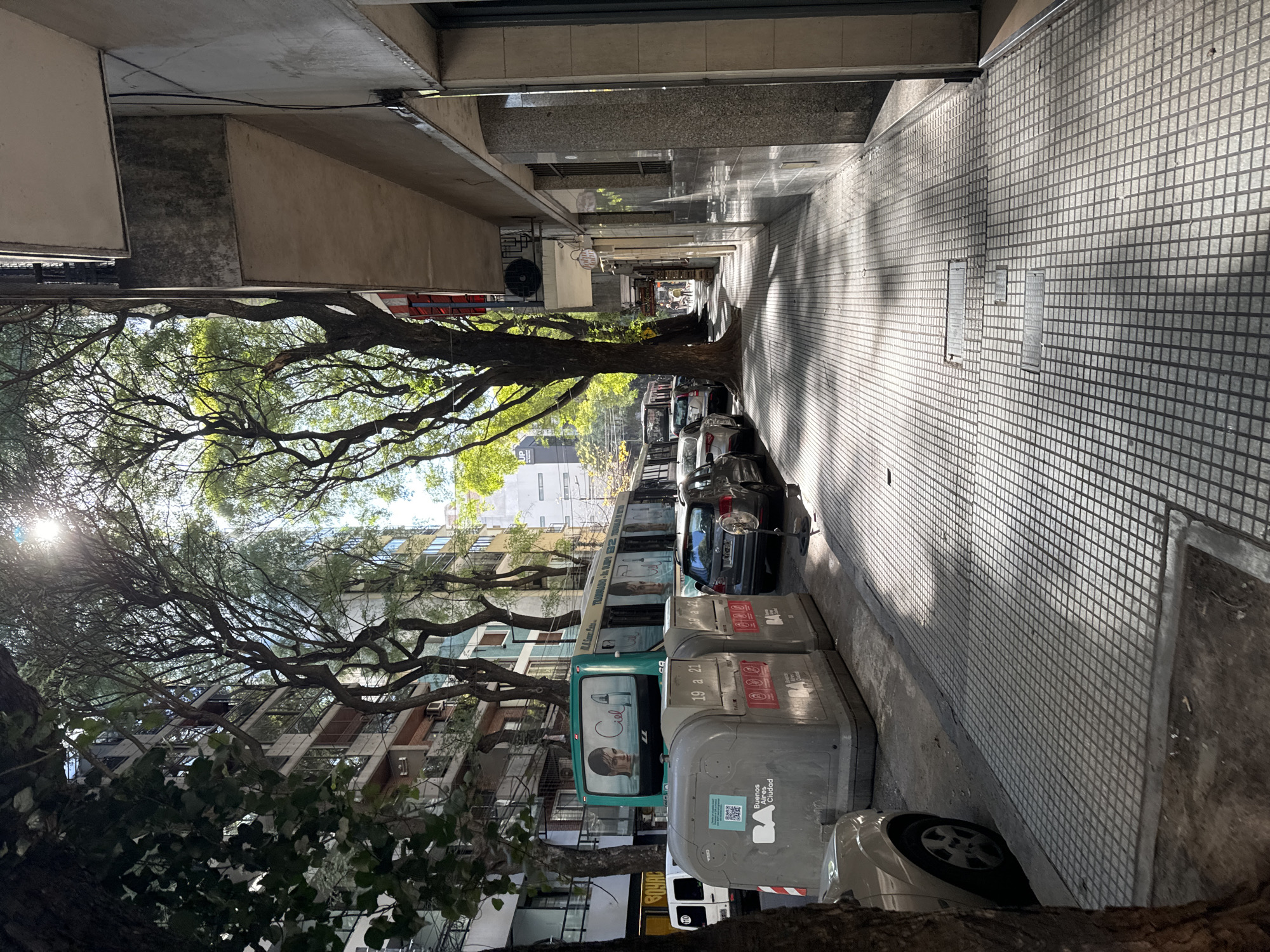 The image shows a street in Buenos Aires, Argentina. The street is lined with buildings on the left and a sidewalk on the right. Trees grow along the street, providing shade. There are cars parked along the street, and trash containers are located on the sidewalk next to the cars. The sidewalk is paved with square tiles. The sun is shining through the trees, creating shadows on the street and sidewalk.