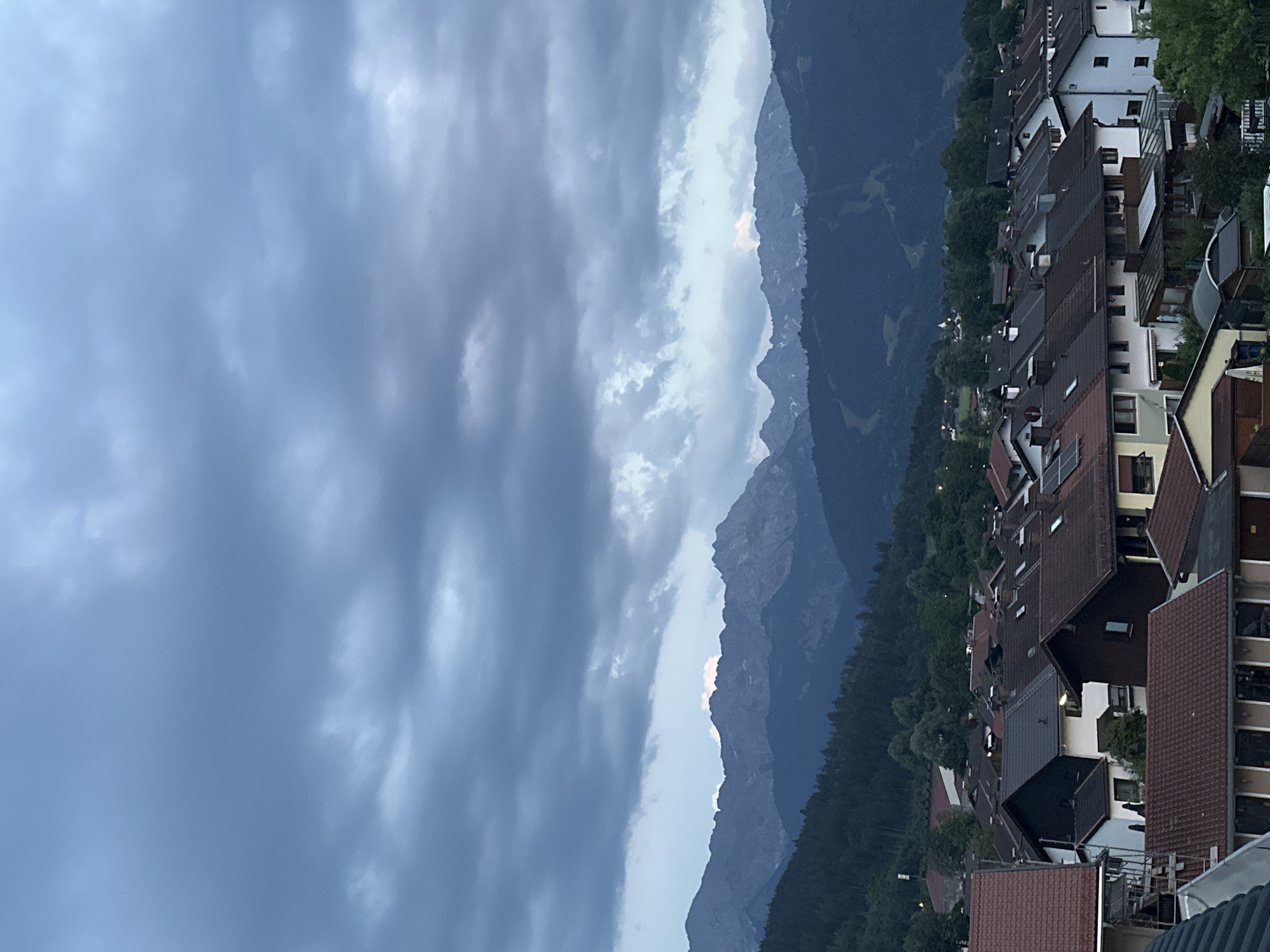 The image captures a twilight view of a residential area in Garmisch-Partenkirchen, Germany, with the Bavarian Alps in the background. The foreground is dominated by a dense cluster of houses with red-tiled roofs, characteristic of alpine architecture. Some houses feature dormer windows and chimneys. The middle ground consists of a dark green forested hillside.
In the distance, a majestic mountain range stretches across the horizon, its peaks softened by the dim light and obscured by a heavy, overcast sky. The clouds are a mix of dark grays and lighter blues, with a subtle hint of pink or orange light breaking through near the mountain peaks, suggesting the last vestiges of sunset or approaching dawn. The overall atmosphere is calm and serene, with a sense of impending evening or early morning. There are no people or animals visible in the frame. The scene appears to be a quiet, residential neighborhood nestled at the foot of the mountains.