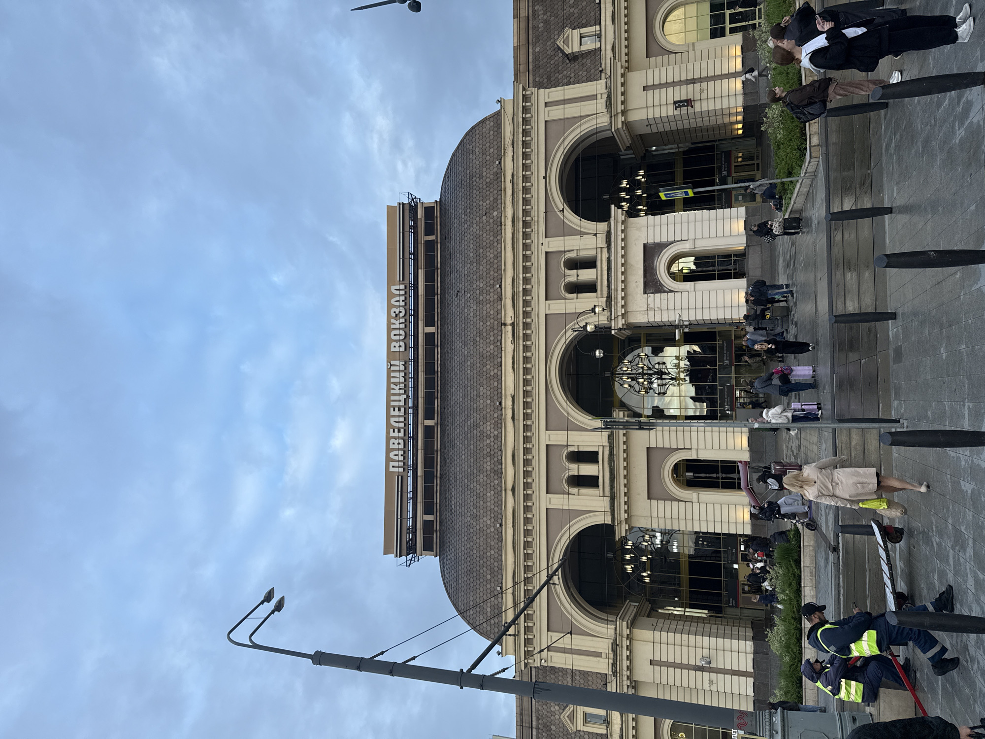 This image shows the Paveletsky railway station (Павелецкий вокзал) in Moscow, Russia. The station building has a light-colored facade with large arched windows and a gray-tiled dome. The name "ПАВЕЛЕЦКИЙ ВОКЗАЛ" is displayed in large letters on top of a section of the building. Several people are walking or standing outside the station, some with luggage. A street lamp is visible in the foreground. Two men in reflective vests are sitting on a metal barrier. The sky is cloudy.