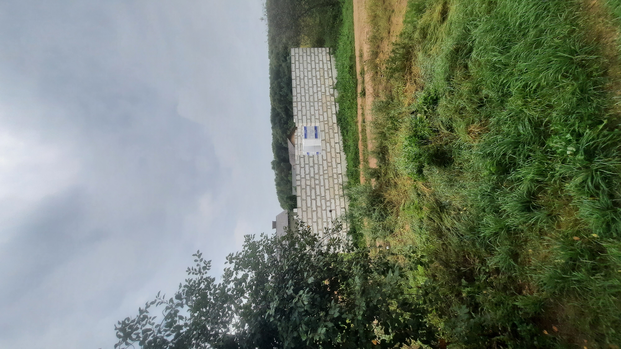 The image depicts a partially constructed cinder block wall in an outdoor environment. The wall is several rows high and appears to be a perimeter boundary. Two stacks of white-wrapped building materials, possibly insulation or more blocks, are visible on top of the wall. The wall is situated in a grassy area with scattered vegetation and a dirt path running in front of it. In the background, a dense line of trees forms a natural horizon, with the roofs of some houses visible peeking through the foliage. The sky is overcast, suggesting a cloudy or possibly rainy day. The overall scene indicates a construction site or a developing property. The location is identified as Shklov, Belarus. There are no people or animals present in the image.